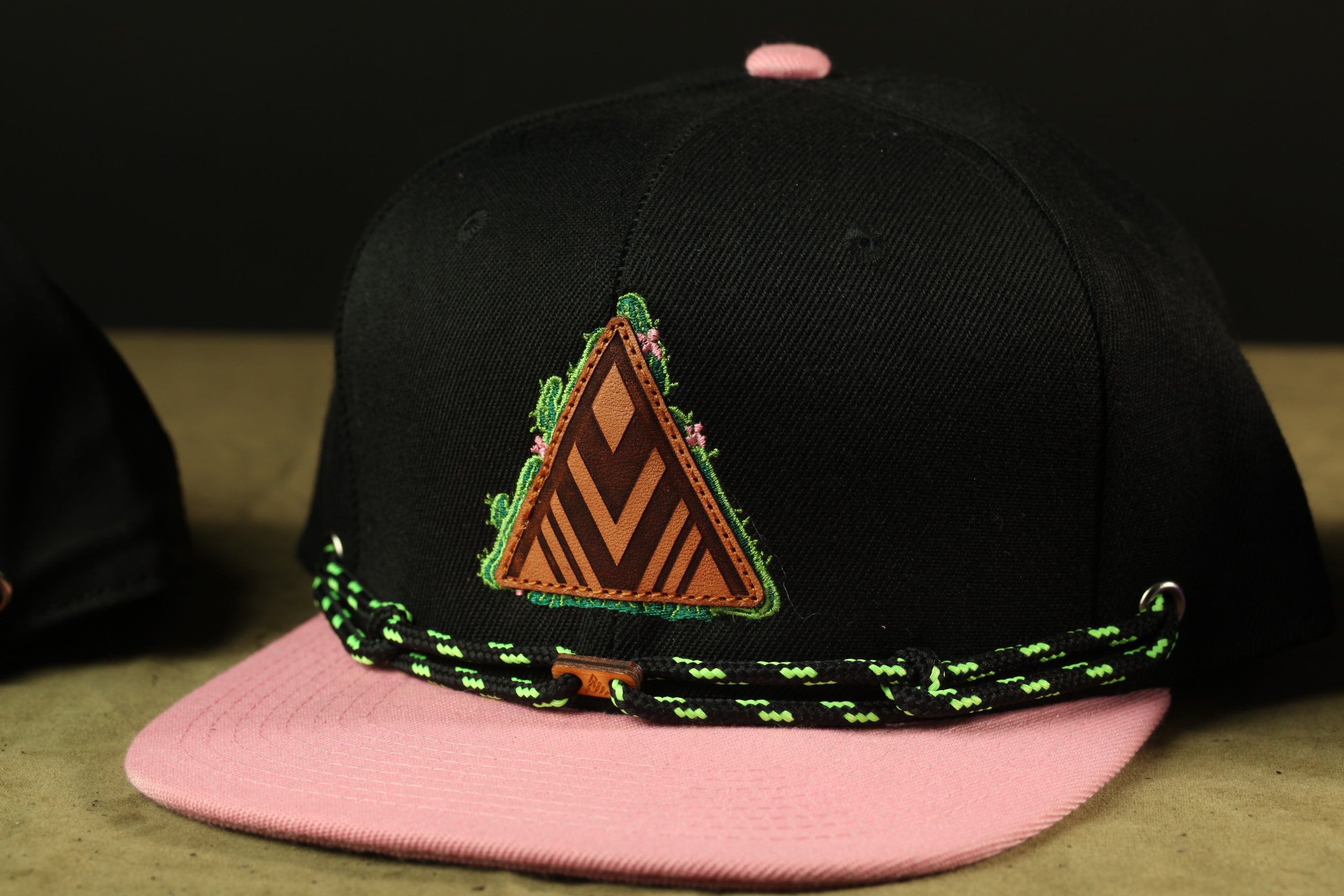 A black baseball cap with a pink brim and a triangular patch featuring a geometric design is shown in the image.