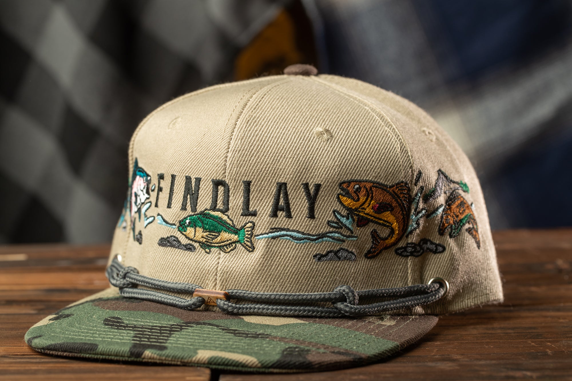 A beige baseball cap with a camouflage pattern and the word "FINDLAY" printed on it, sitting on a wooden surface.