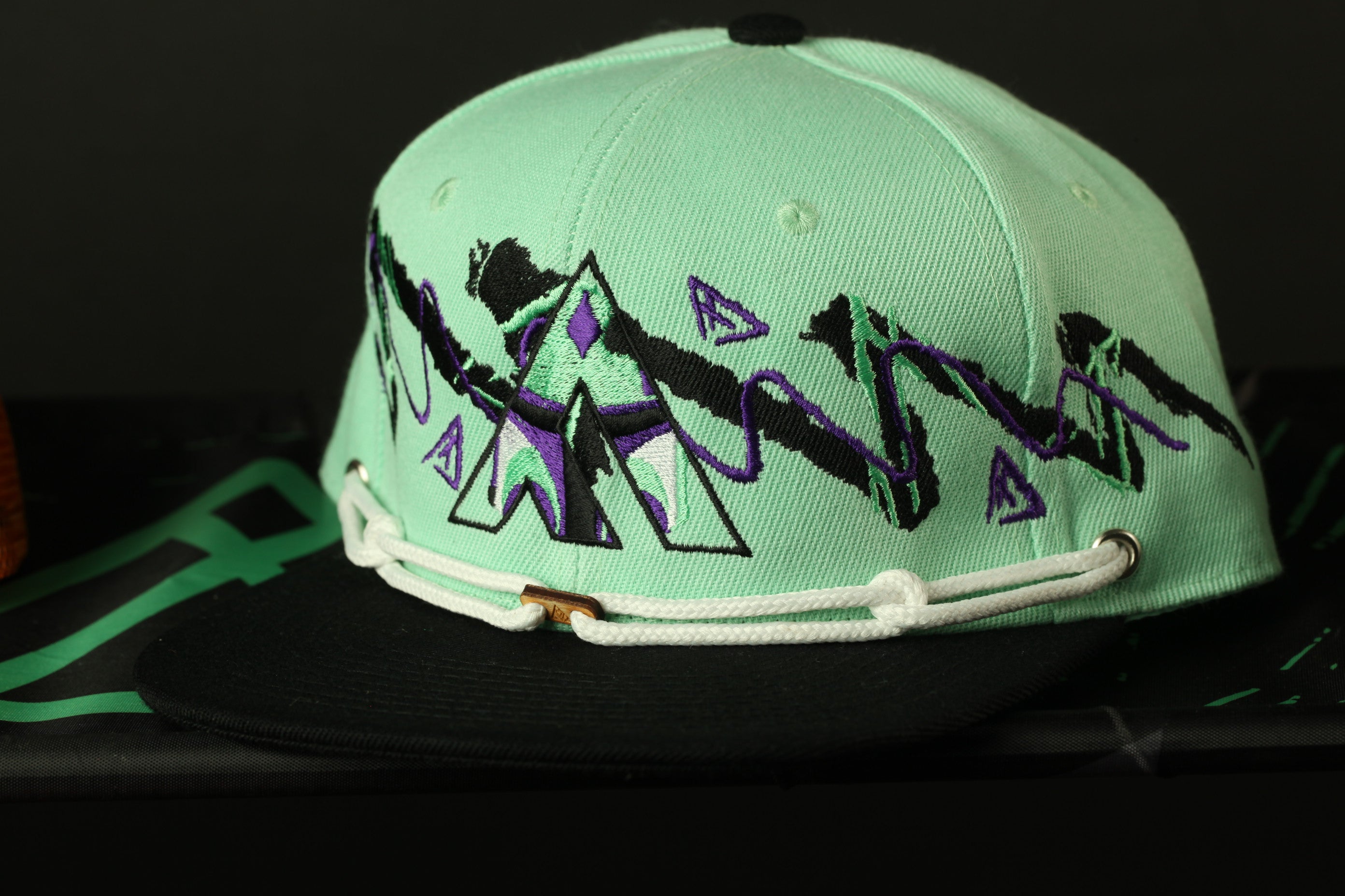 A turquoise baseball cap with a graphic design on the front, featuring purple and green elements.