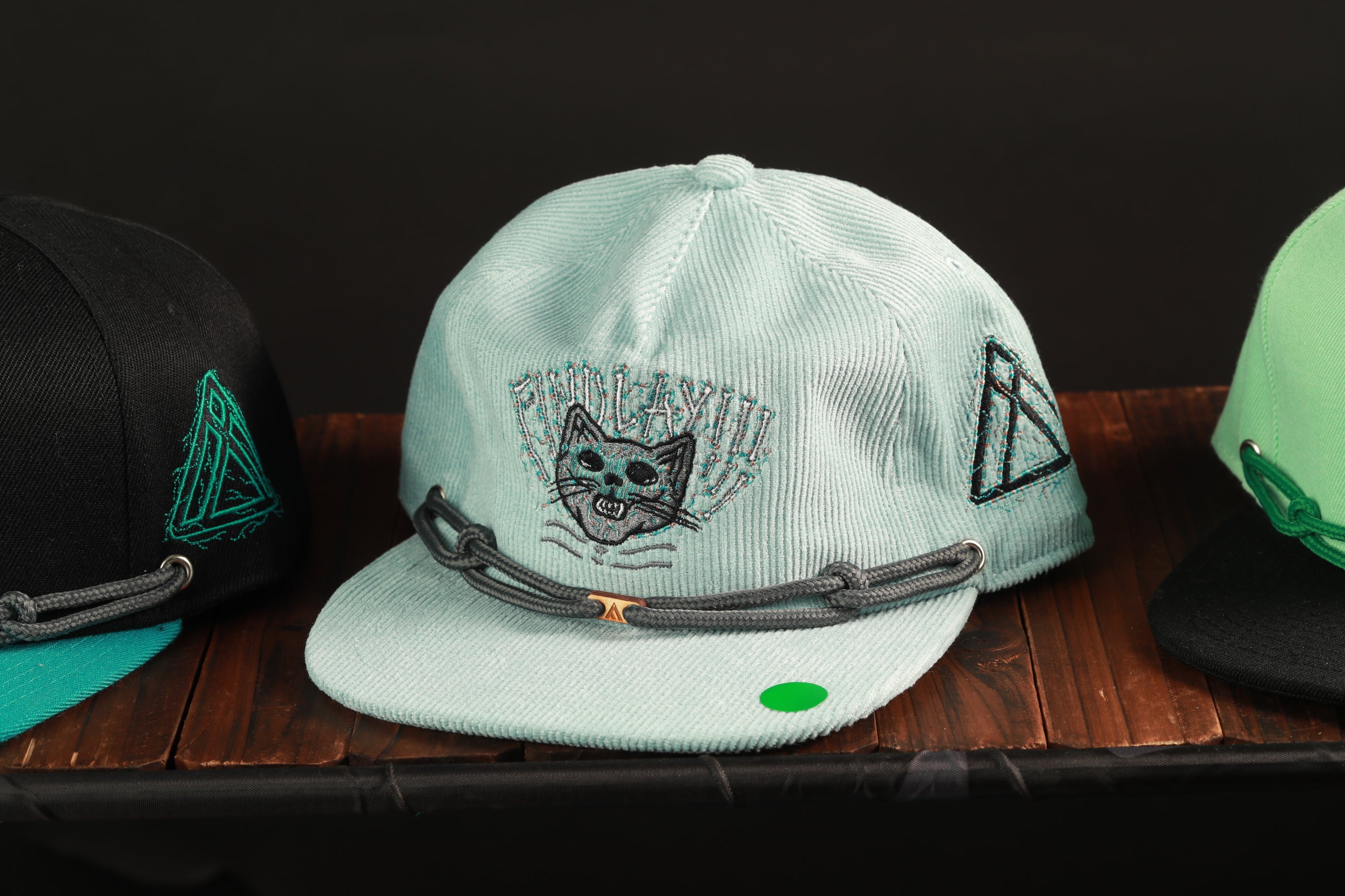 A light blue baseball cap with a cat design and a green tag is displayed on a wooden surface, accompanied by two other baseball caps in the background.