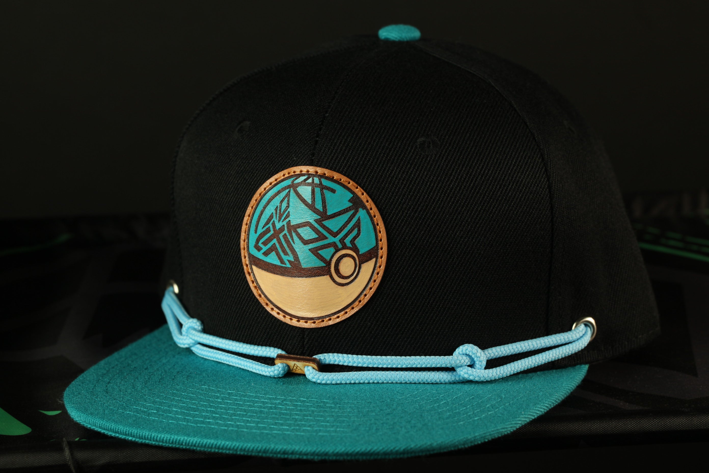 A black baseball cap with a teal brim and a circular logo on the front, resting on a black surface.