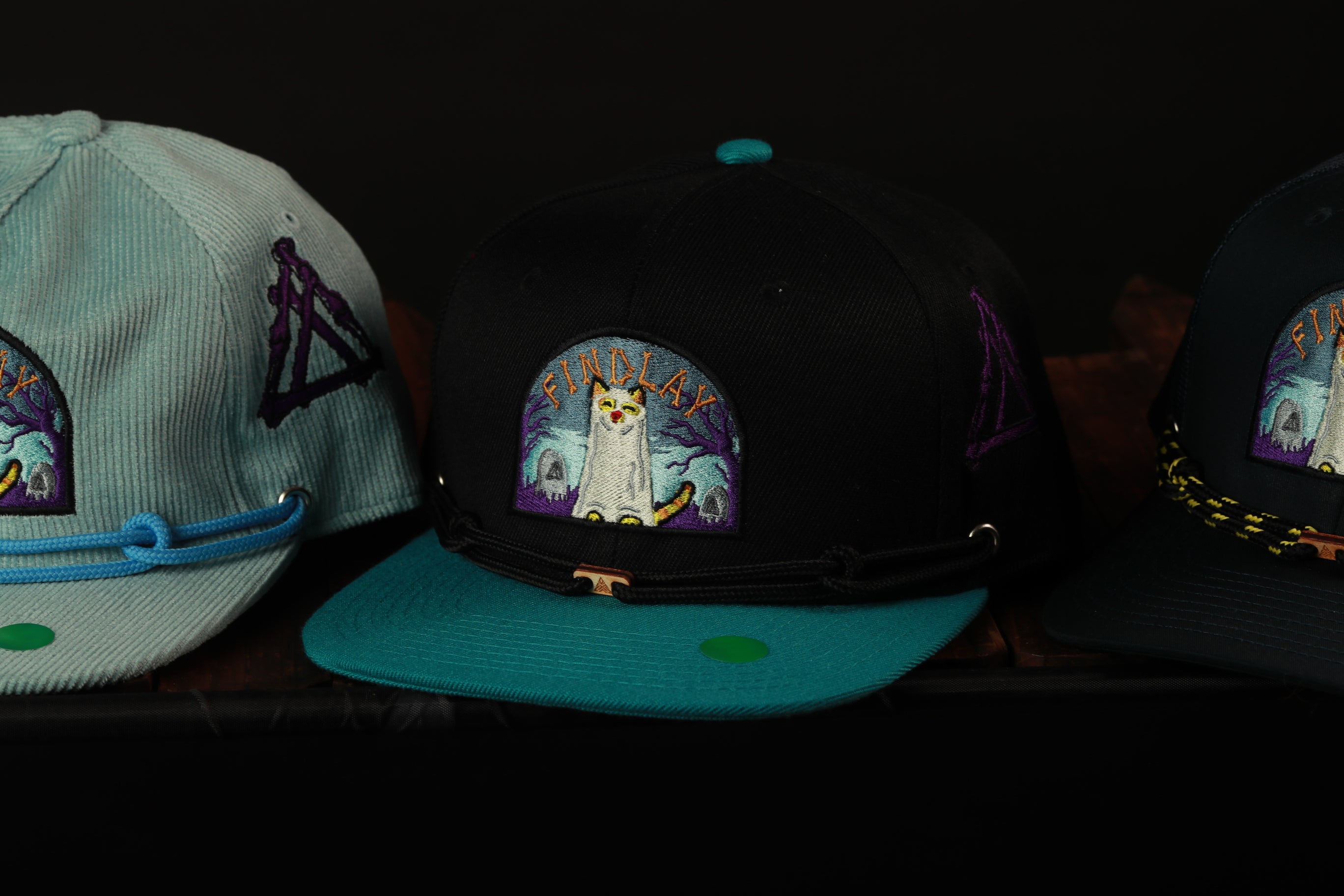 Three baseball caps with different colors and designs, including a black cap with a cat design and two other caps in shades of blue and black.