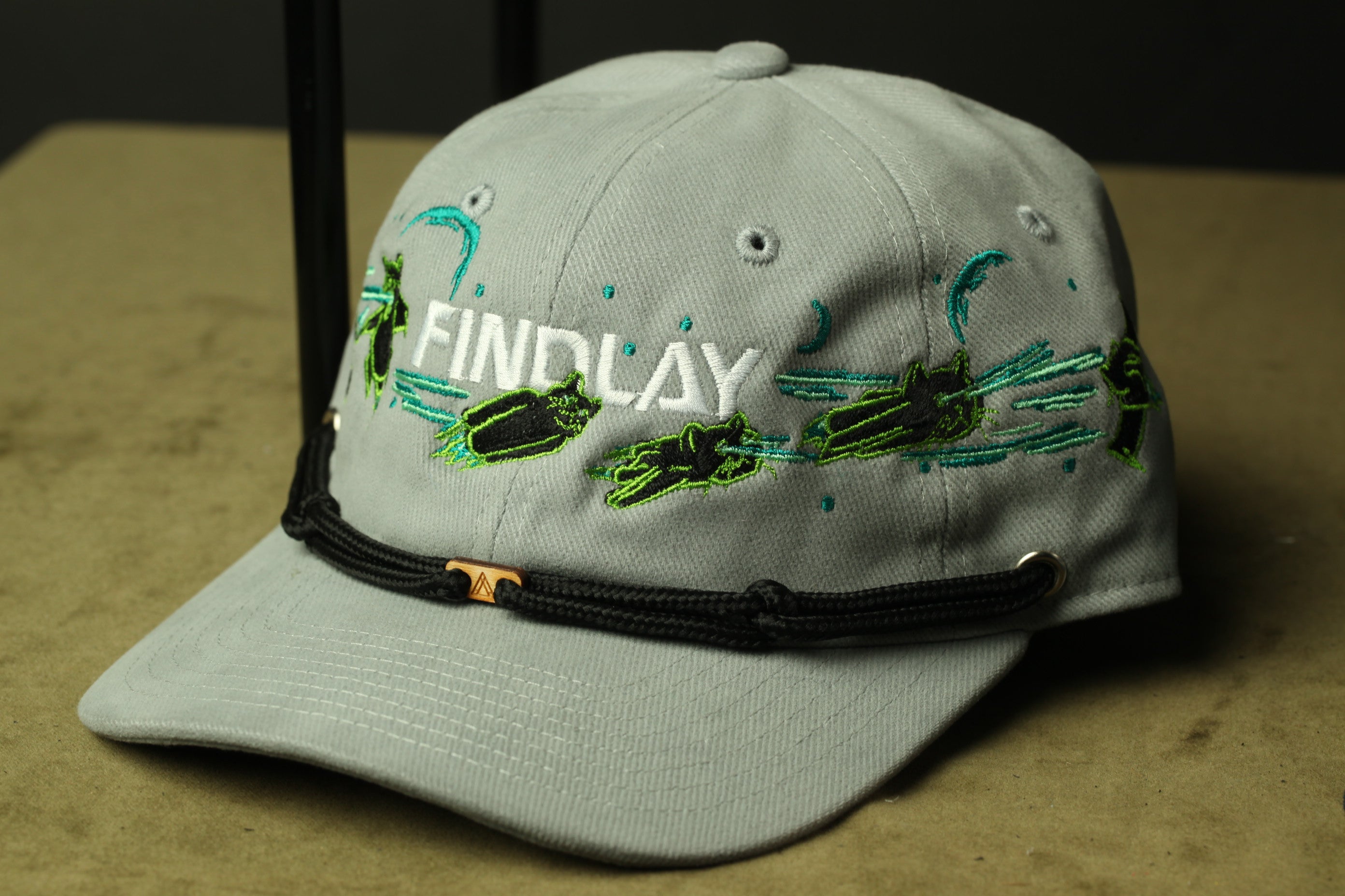 A gray baseball cap with the word "FINDlay" embroidered on it, along with various green and black designs.