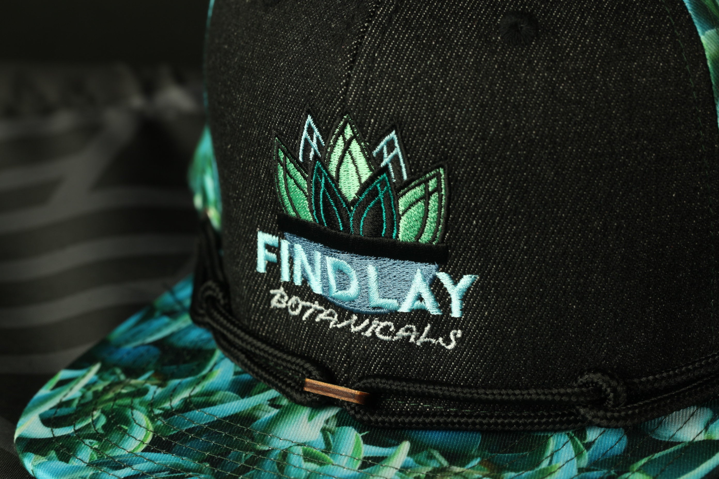 A close-up of a black baseball cap with a floral pattern and the text "FINDLAY BOTANICALS" printed on it.