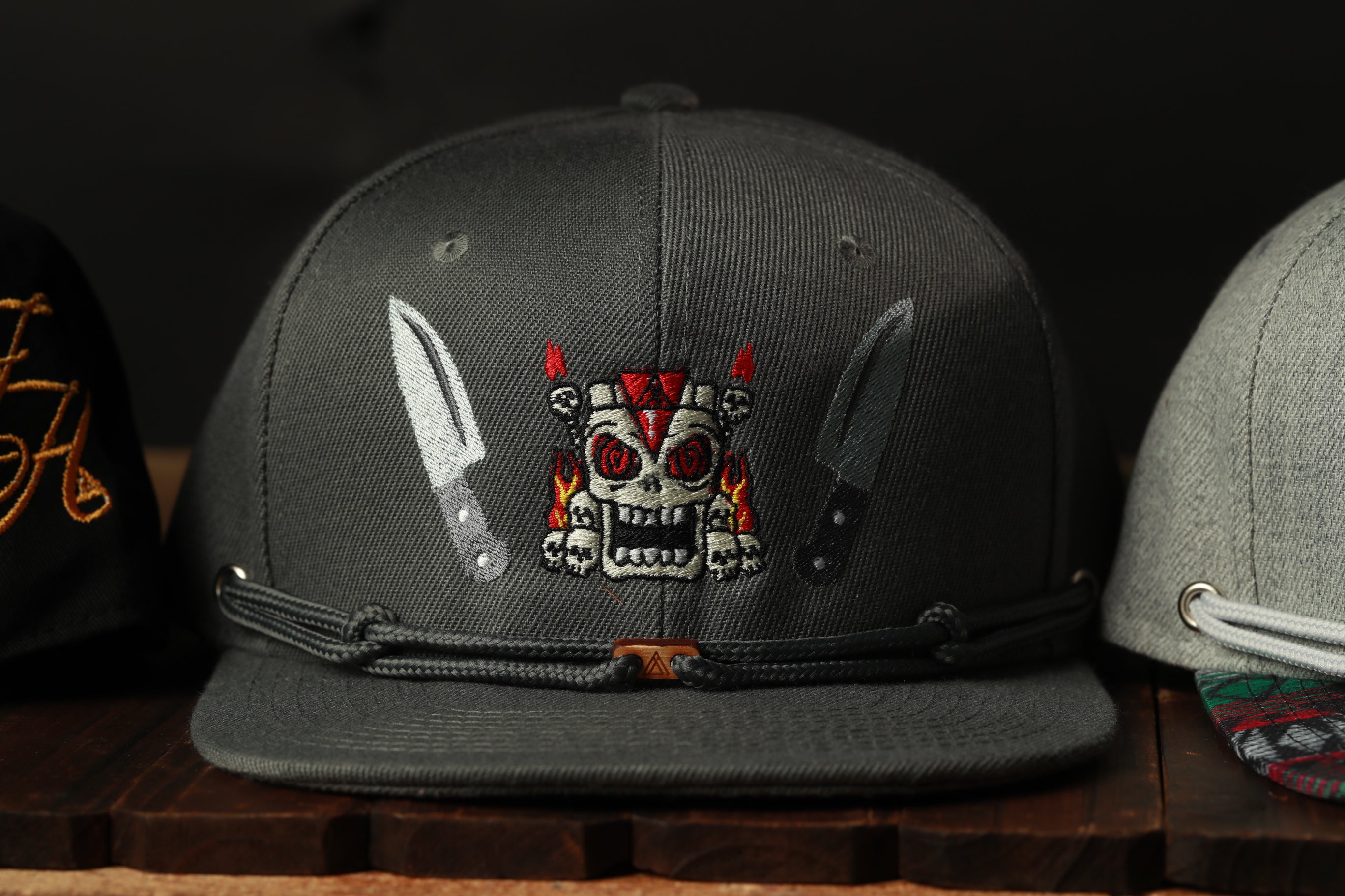 A gray baseball cap with a skull and crossbones design on the front, along with two knives on either side of the skull.