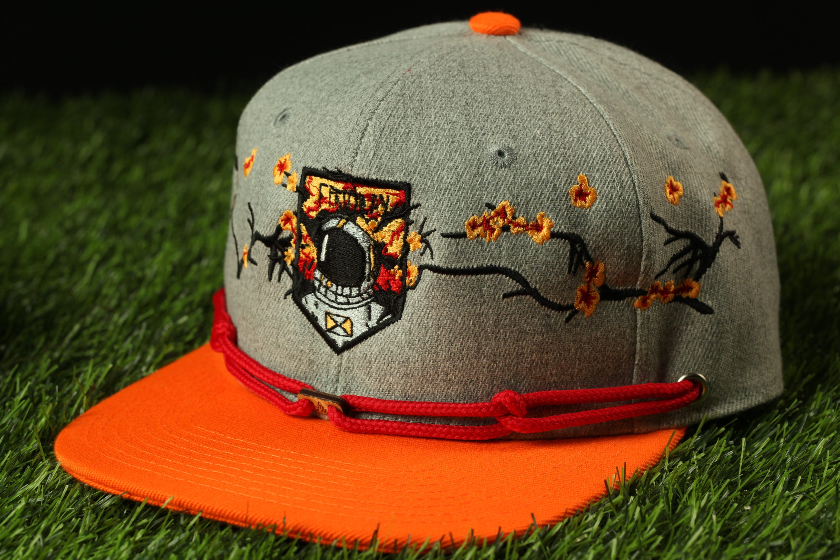 A gray baseball cap with an orange brim and a red string, featuring a black and white emblem with a skull and crossbones design.