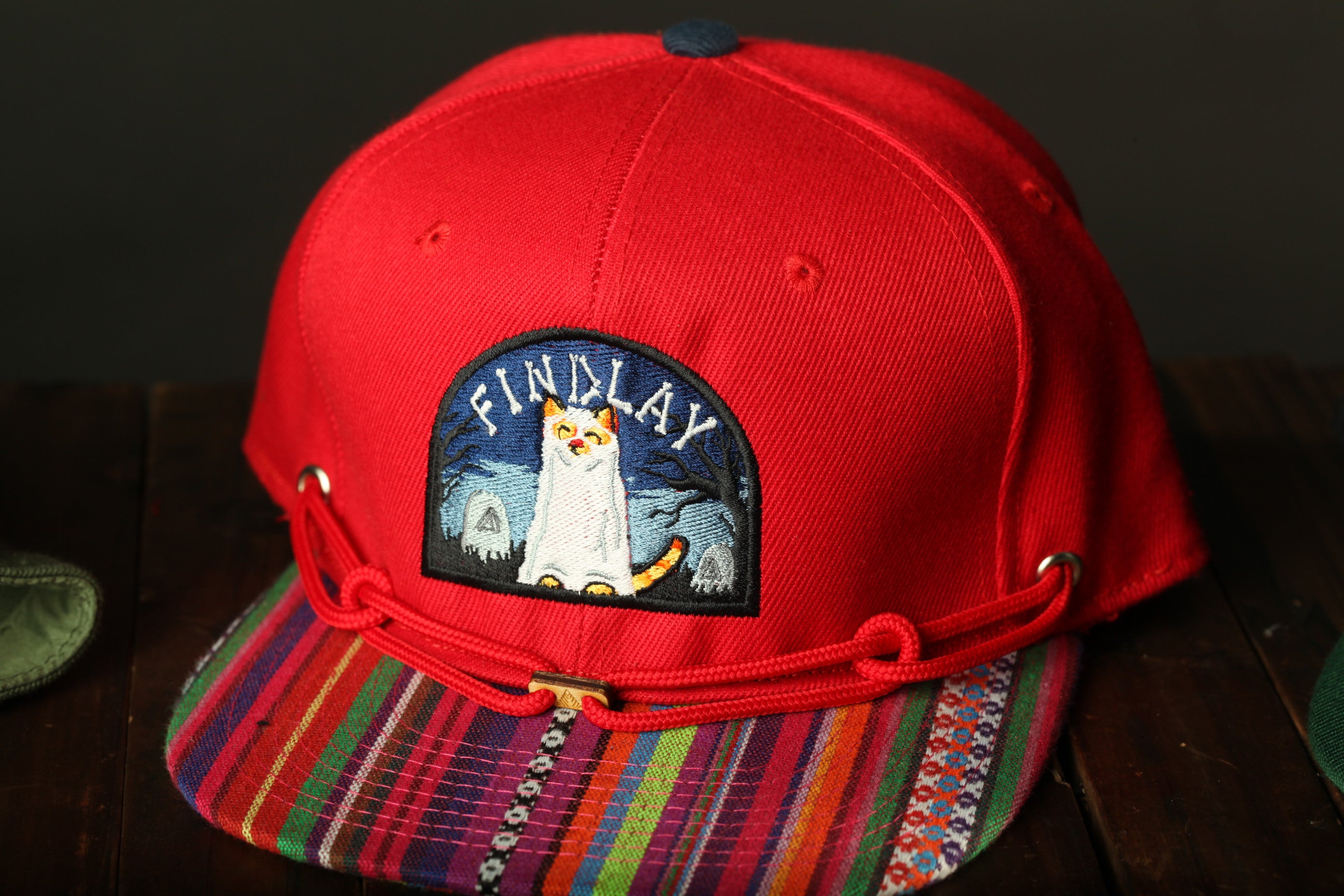 A red baseball cap with a colorful, multicolored brim and a patch featuring a cat design.
