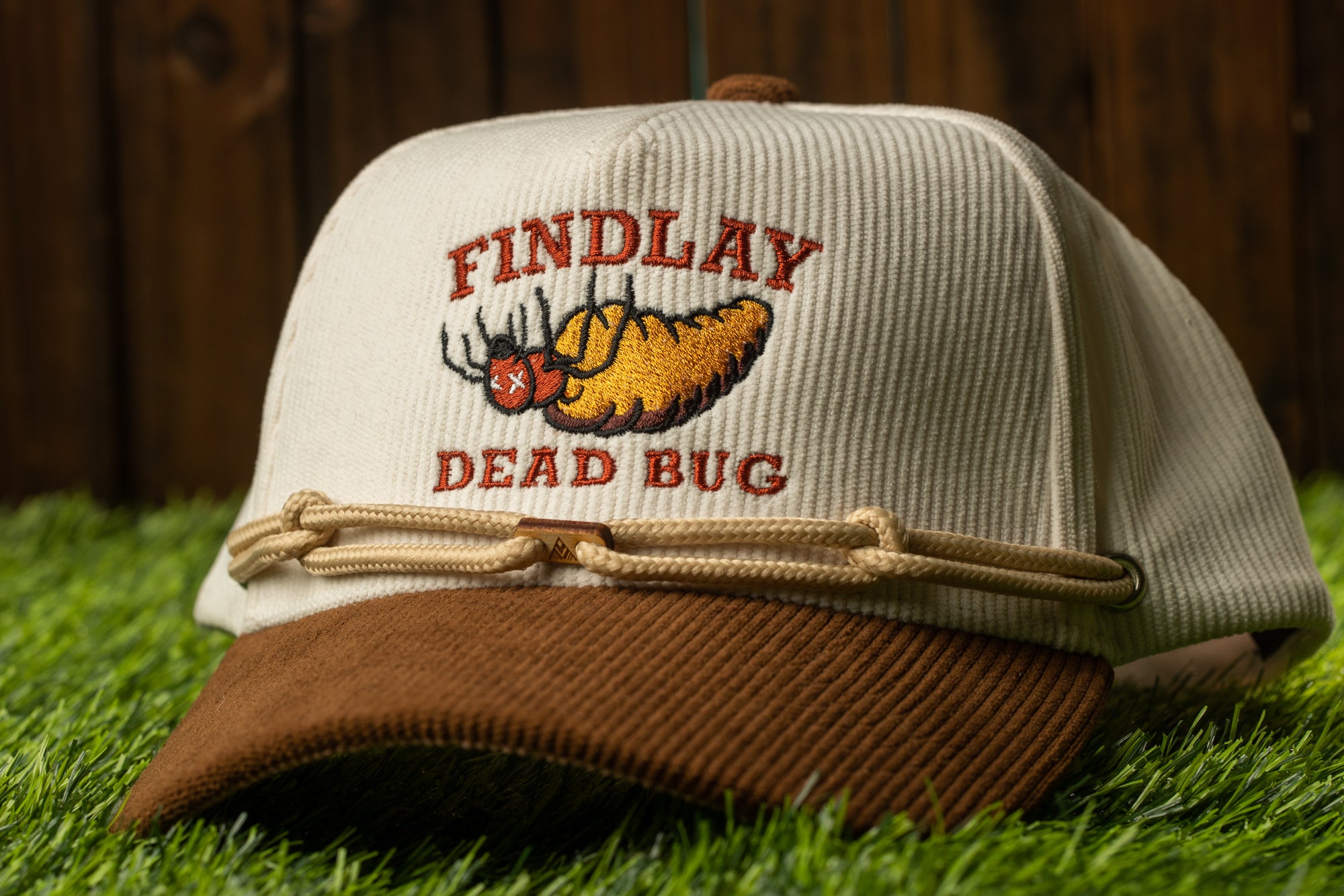 A beige baseball cap with a brown brim, featuring a cartoon image of a dead bug and the text "FINDLAY DEAD BUG" printed on it.