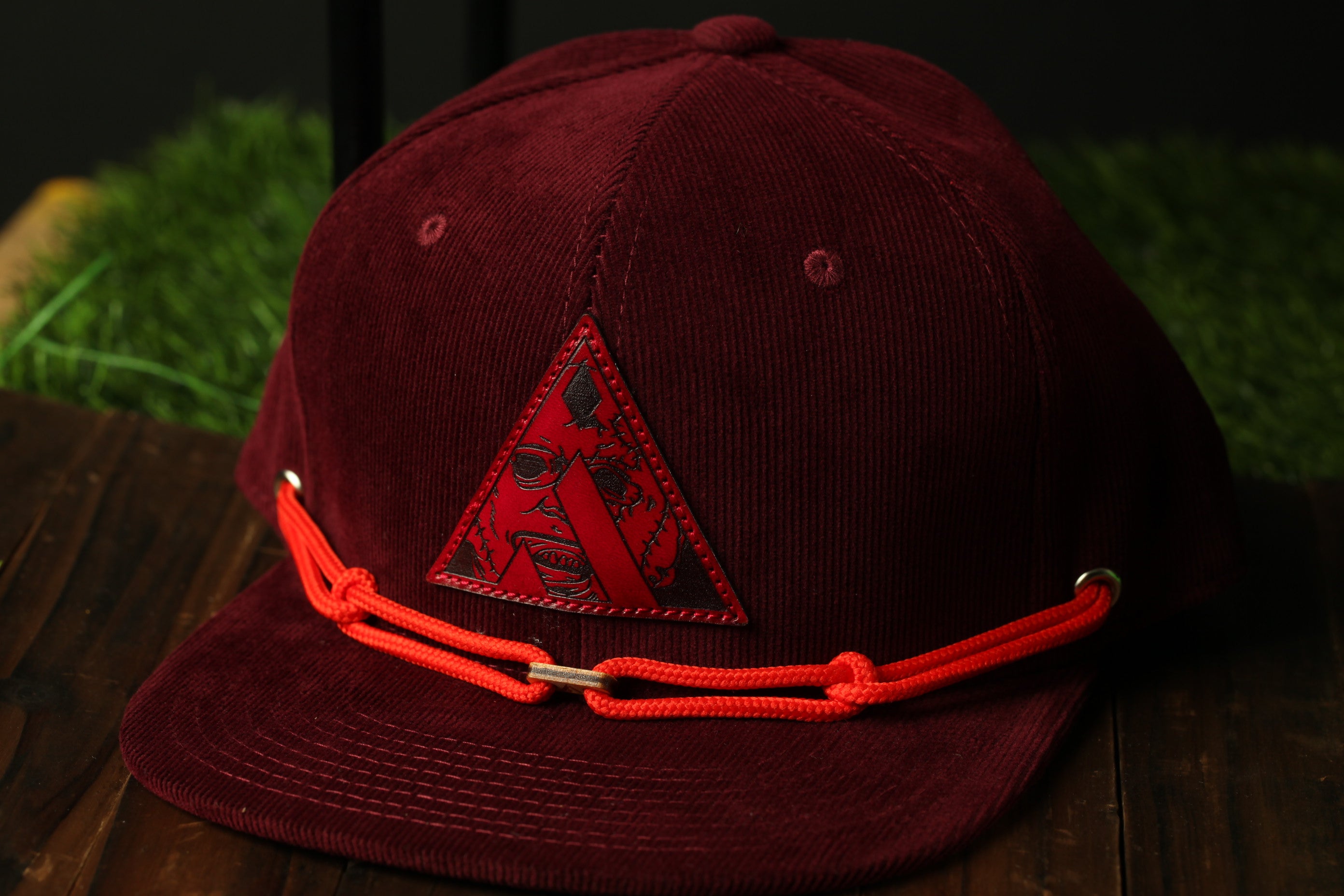 A burgundy baseball cap with a red triangle logo and orange straps is displayed on a wooden surface.