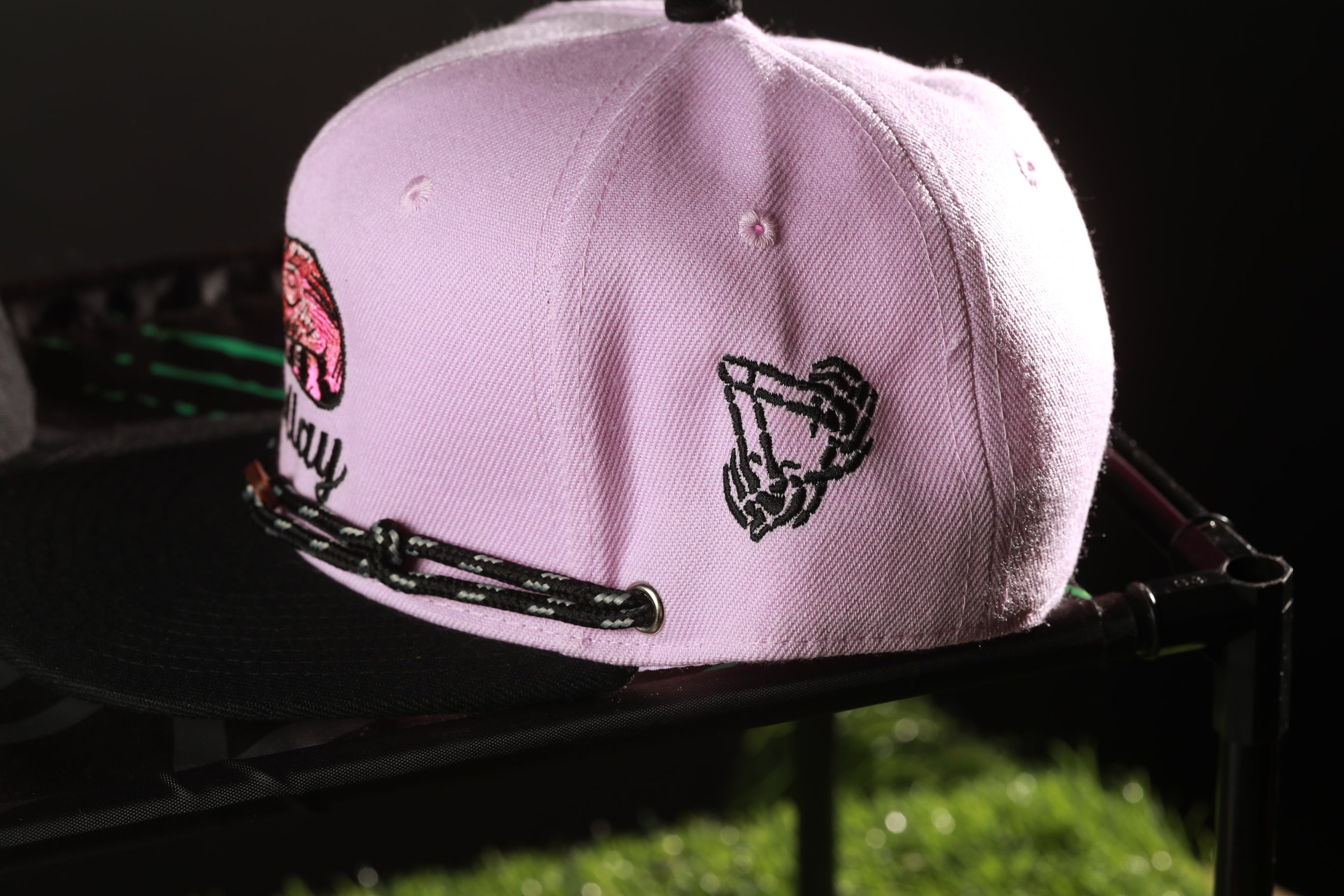 A pink baseball cap with a black graphic design on the side, placed on a black surface.
