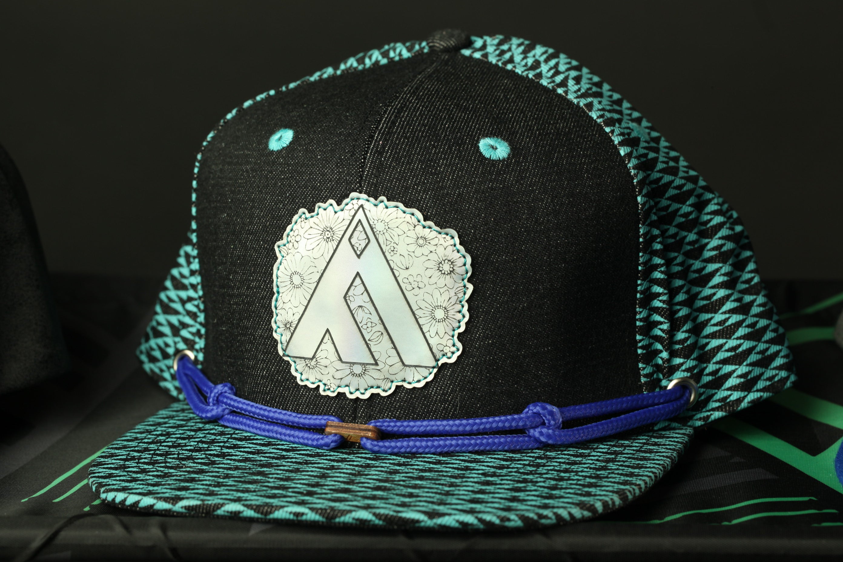A black baseball cap with a teal and black patterned brim, featuring a circular logo on the front.