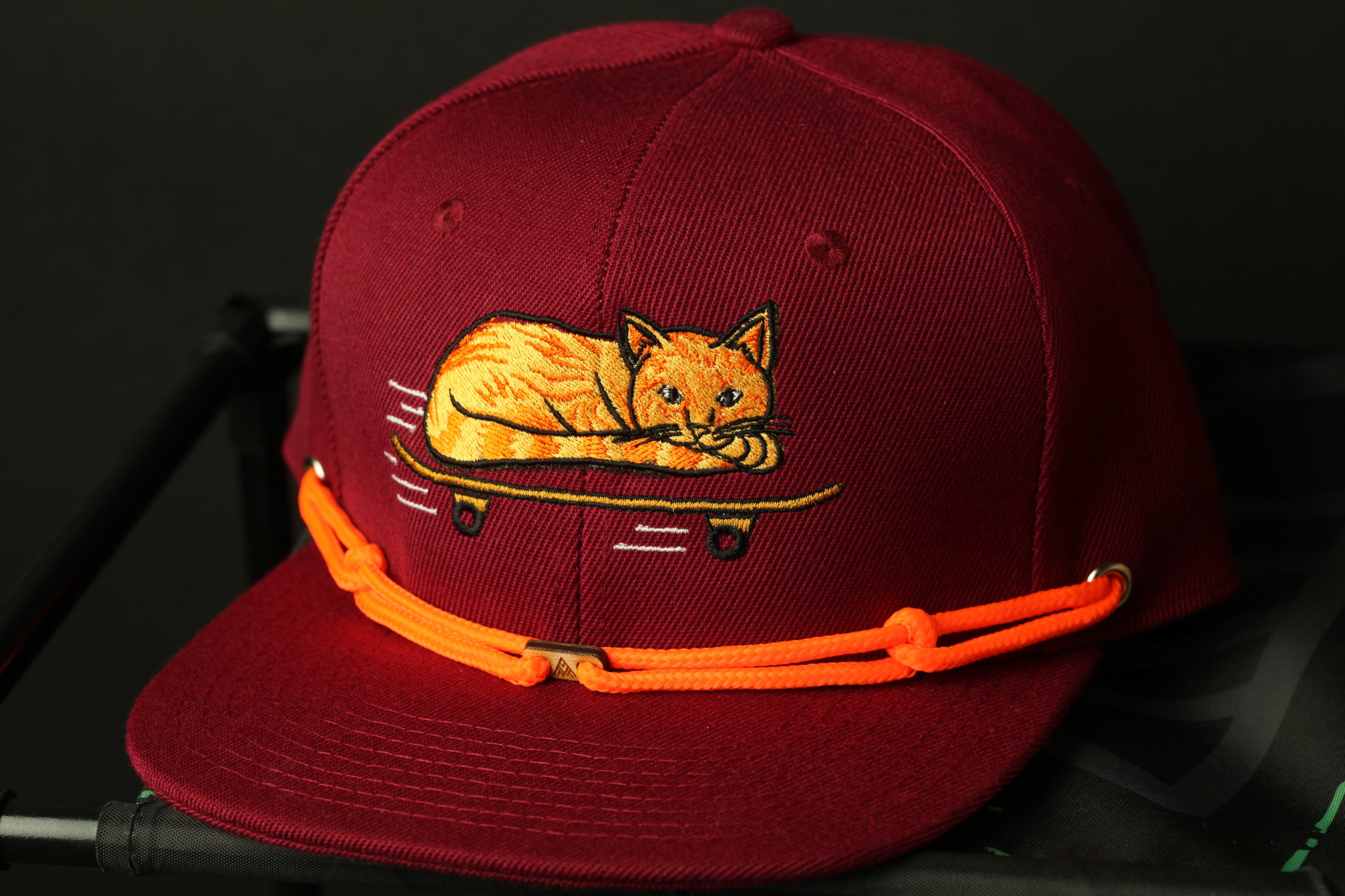 A maroon baseball cap with an embroidered image of a cat riding a skateboard on it.