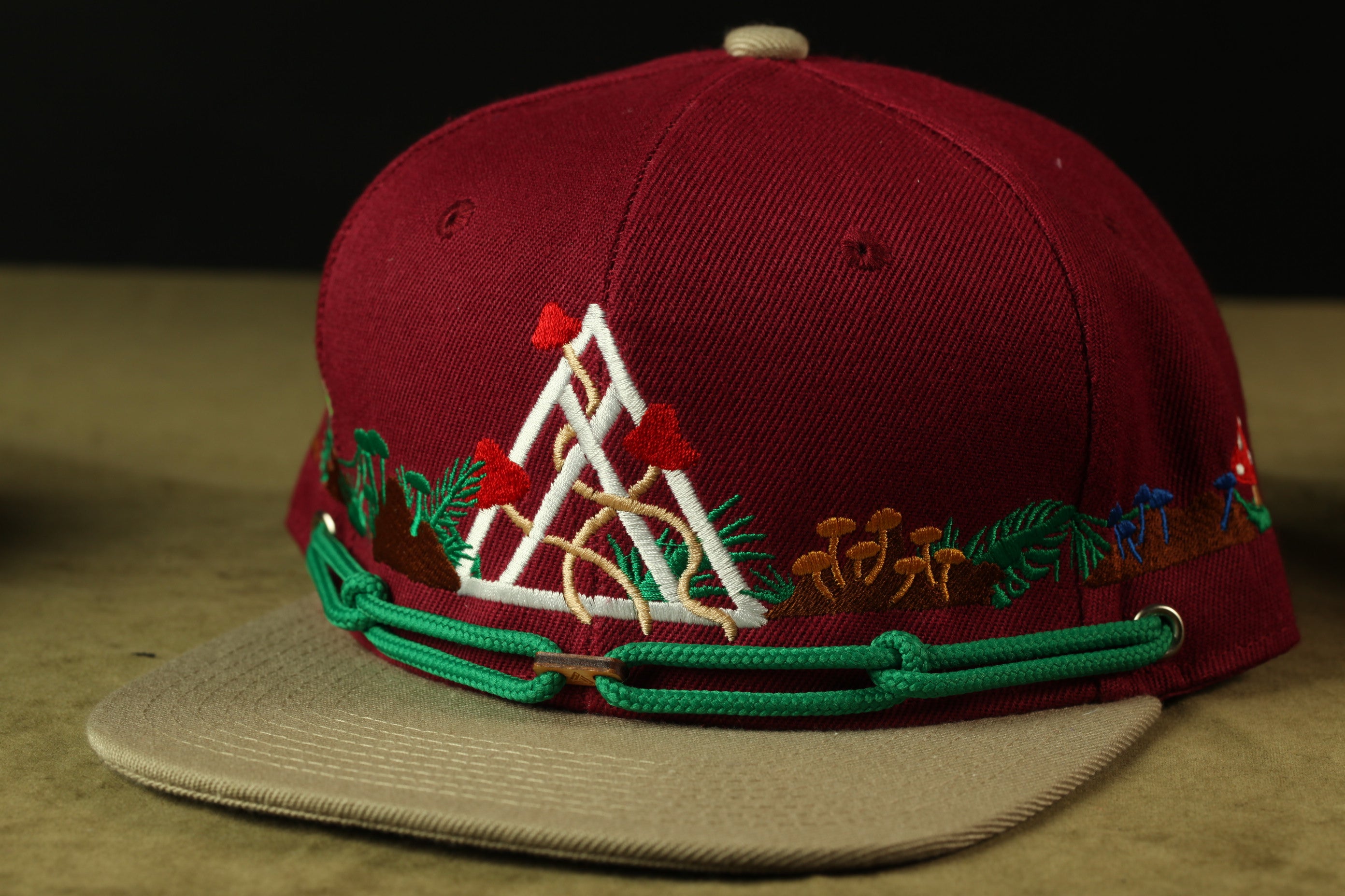 A maroon baseball cap with a green and white embroidered design, featuring a triangle, flowers, and mushrooms, is shown in the image.