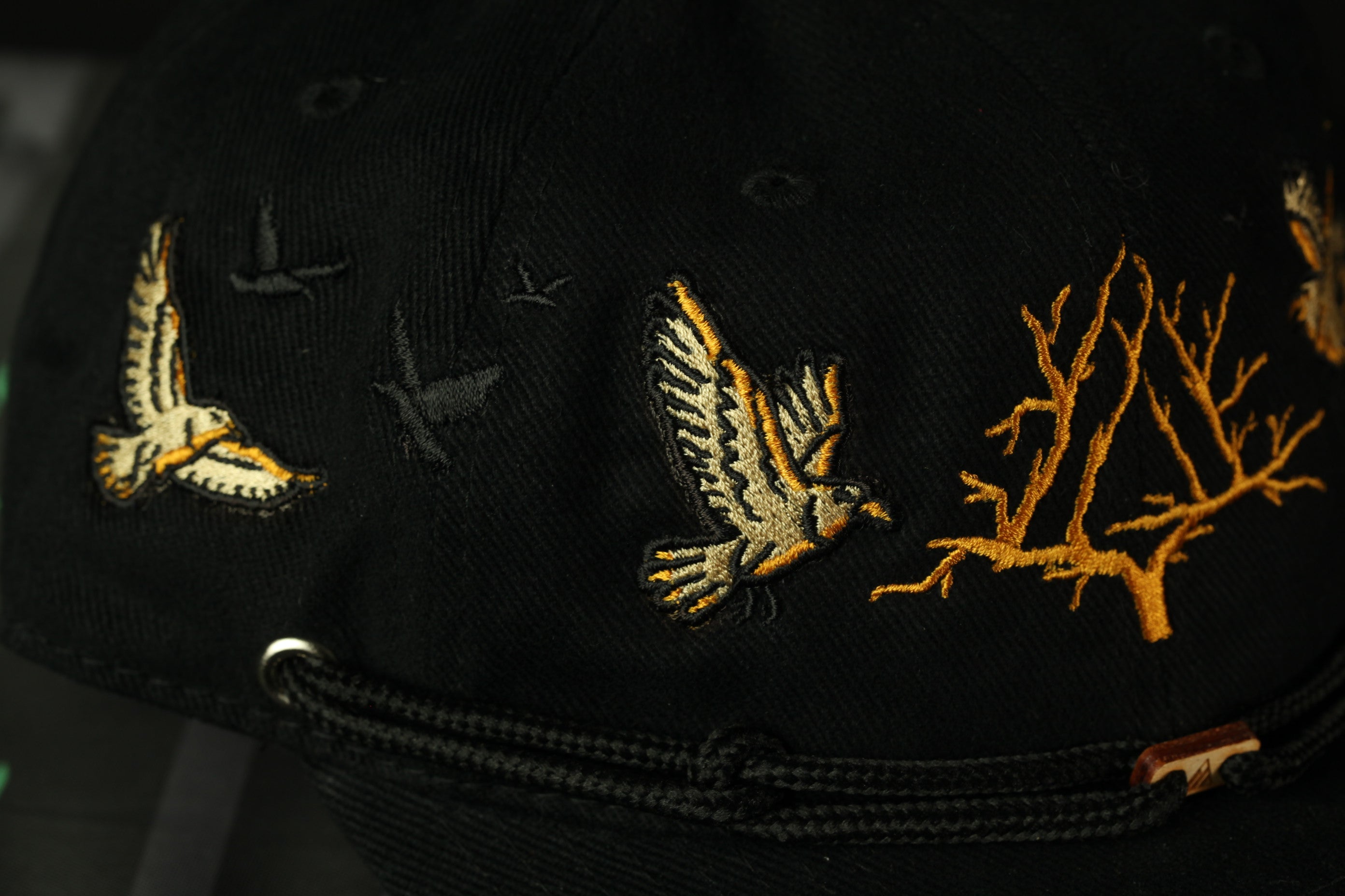 A close-up view of a black baseball cap with gold embroidered designs, including a bird and a tree branch.