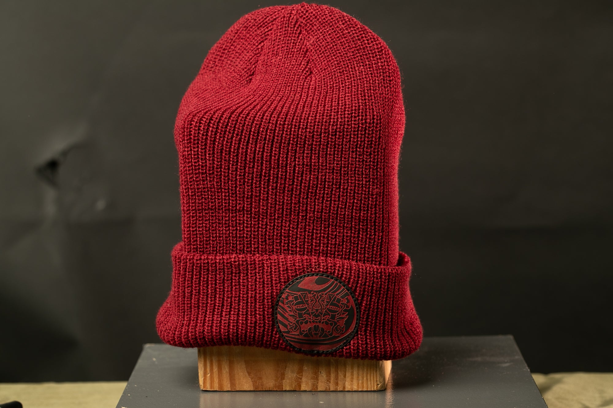 A red knit beanie with a black circular patch on the front, displayed on a wooden stand against a dark background.