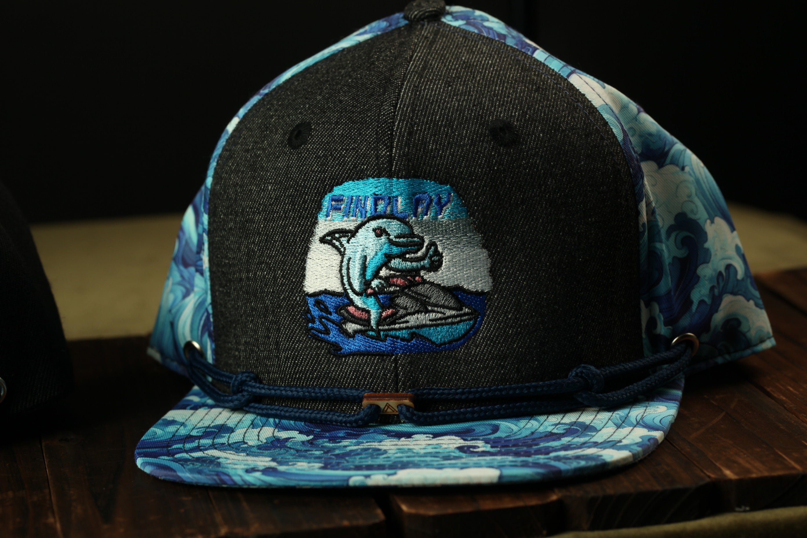 A black baseball cap with a blue and white wave pattern and a graphic of a dolphin on the front.