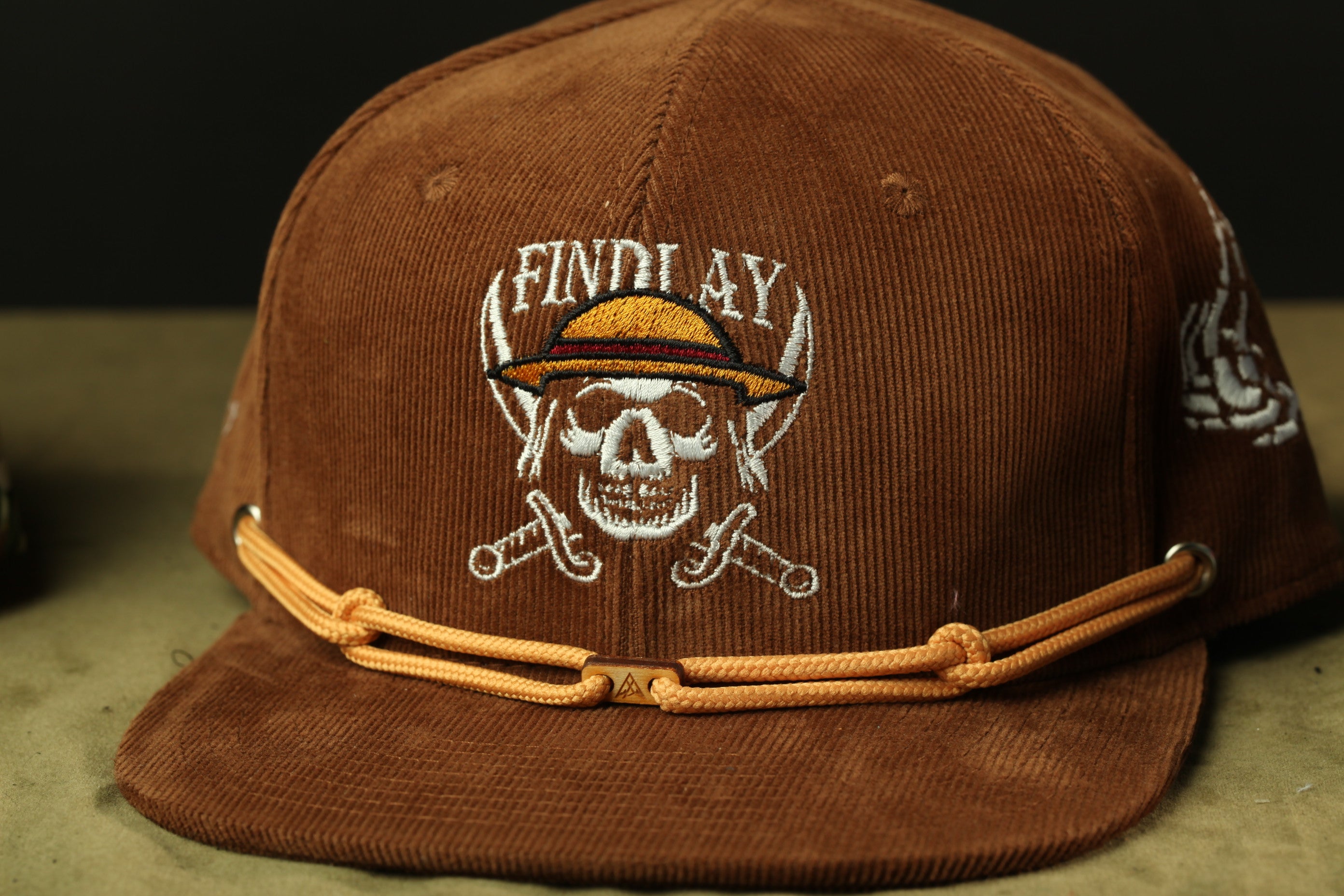 A brown baseball cap with a skull and crossed swords design, and the word "FINDlay" printed on it.