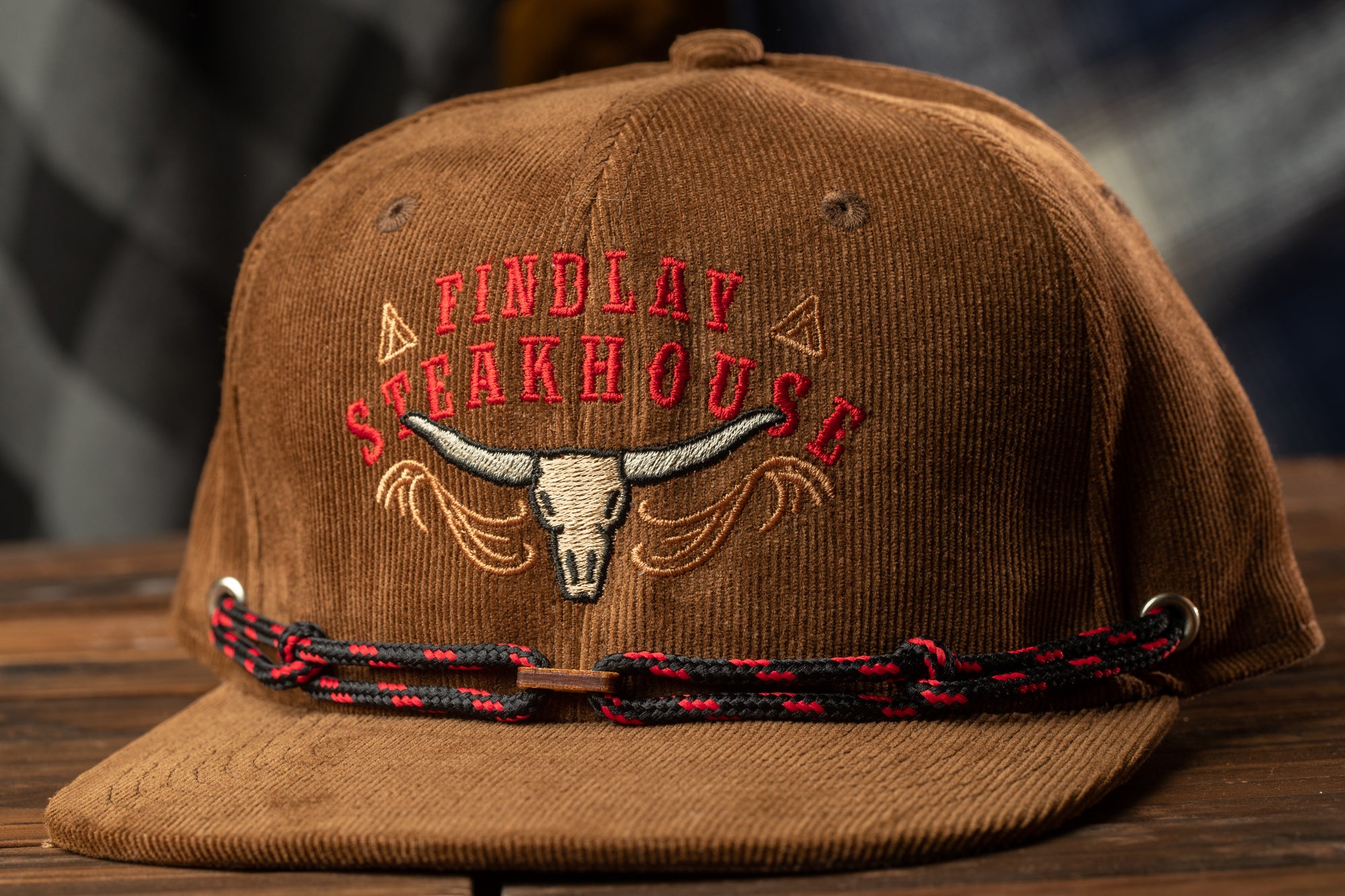 A brown baseball cap with a red and black braided strap and embroidered text on the front, which says "Findlay Steakhouse".