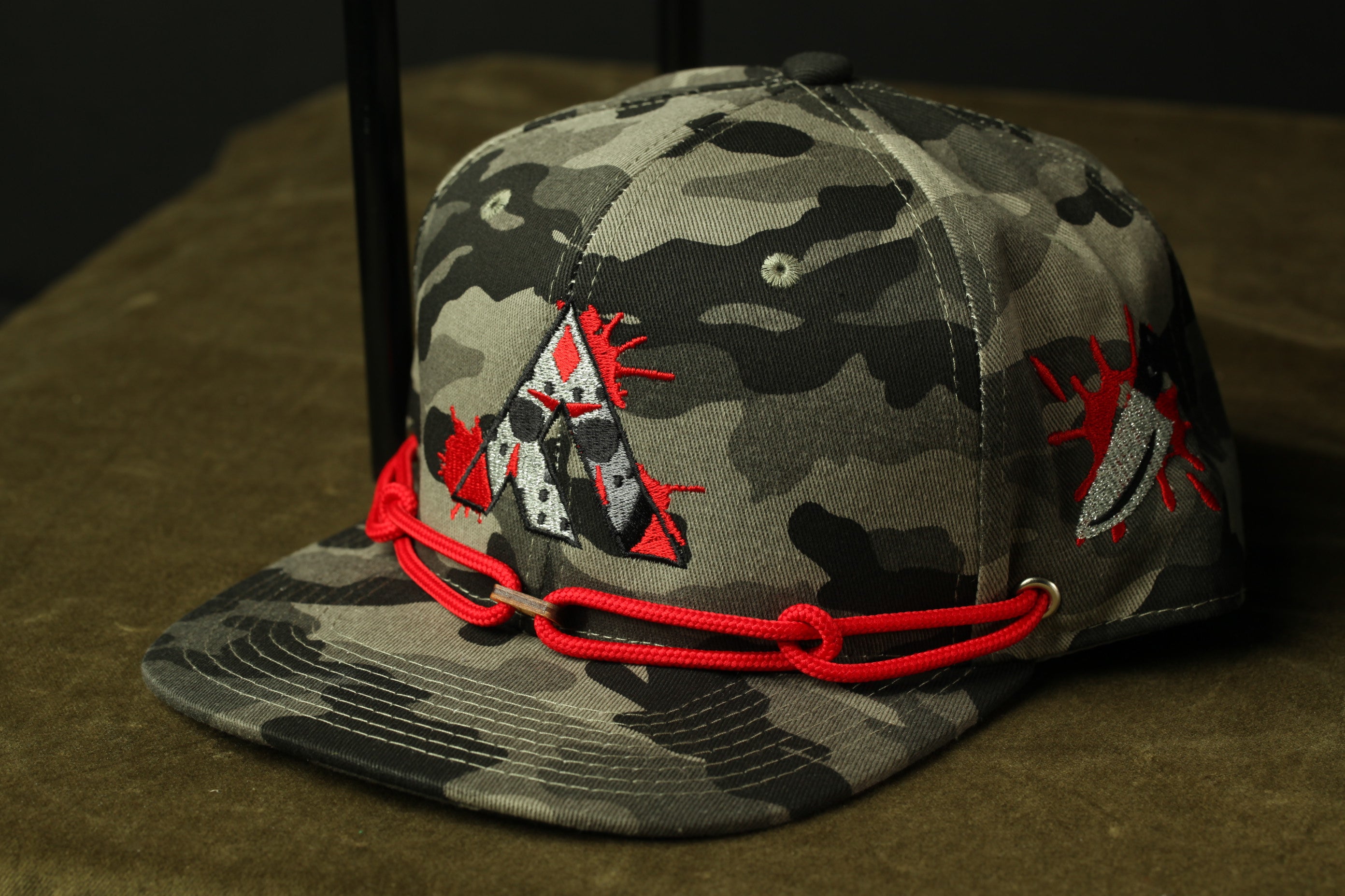 A camouflage baseball cap with a red rope chain and a graphic design on the front.