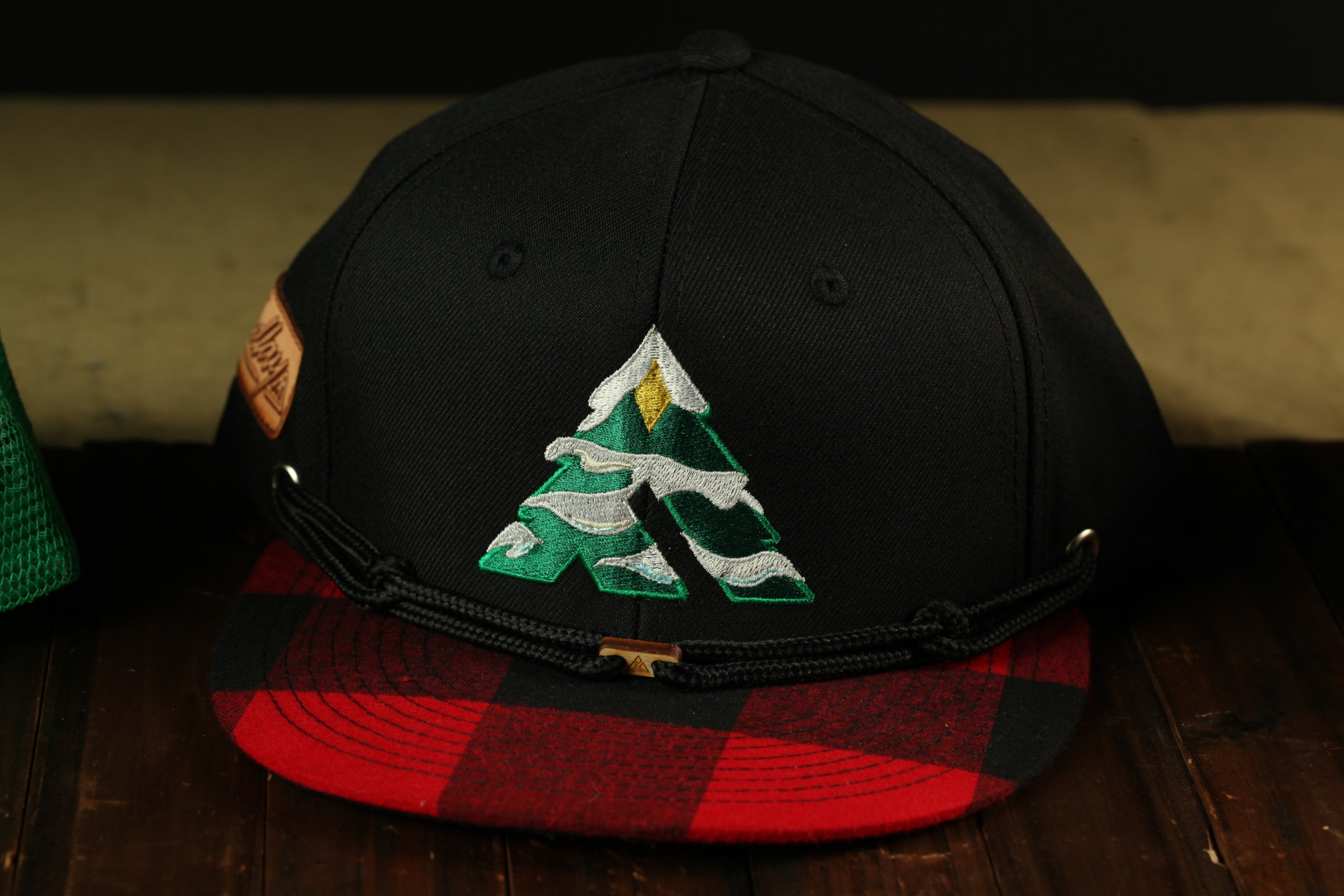 A black baseball cap with a red and black plaid pattern and a green tree embroidered on the front.