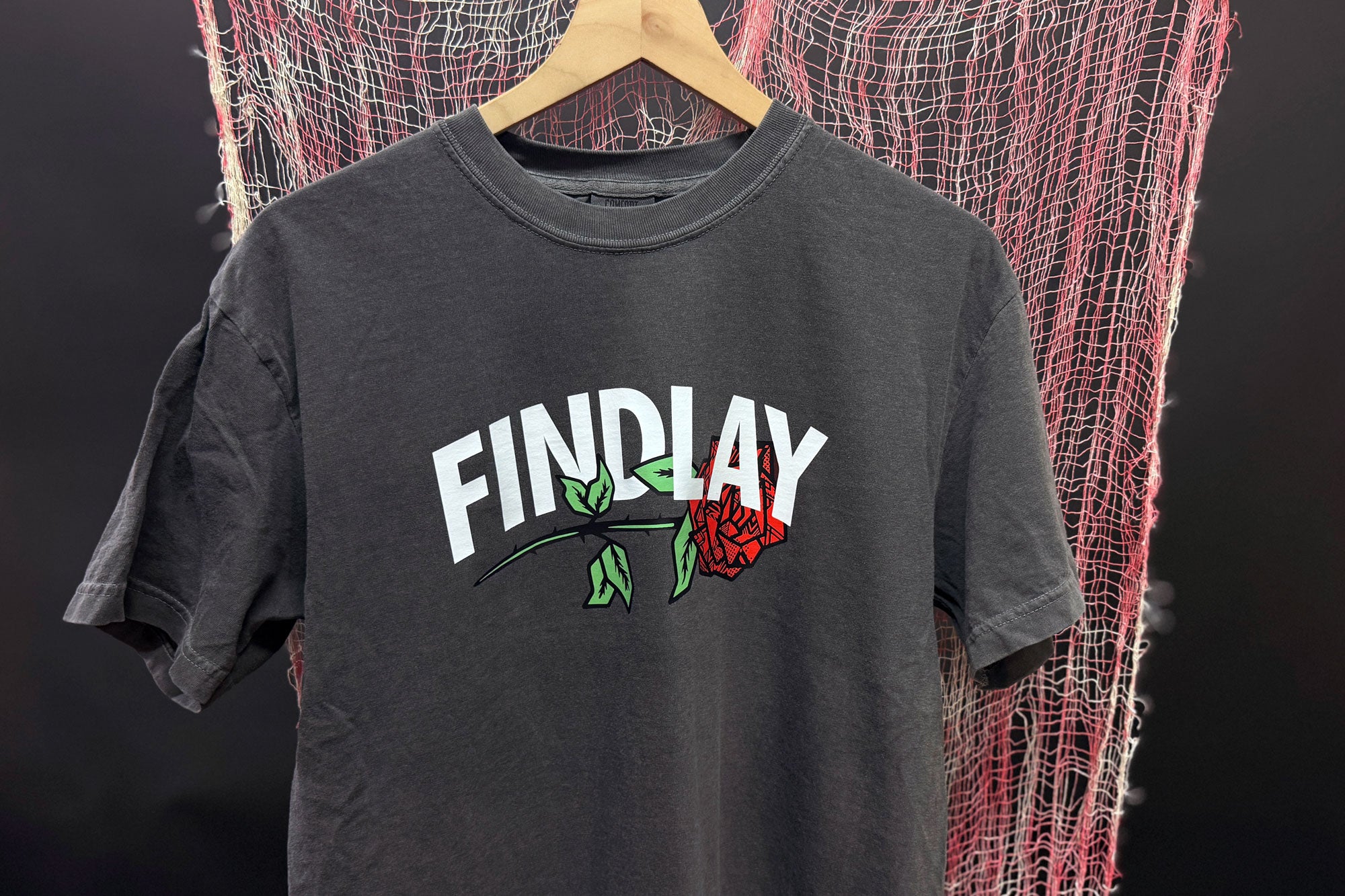 A gray t-shirt with the word "FINDlay" printed on it, hanging on a wooden hanger against a background of red and white netting.