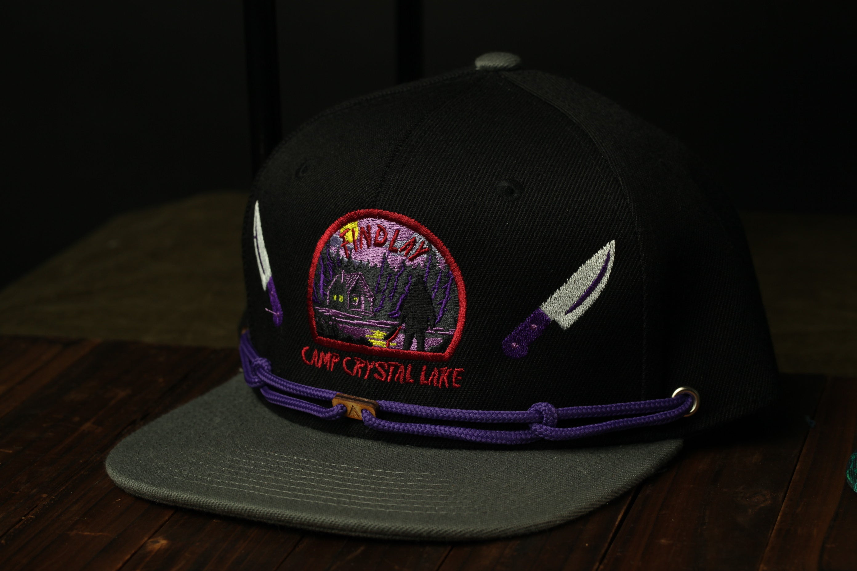 A black baseball cap with purple and white accents, featuring an embroidered logo and text, placed on a wooden surface.