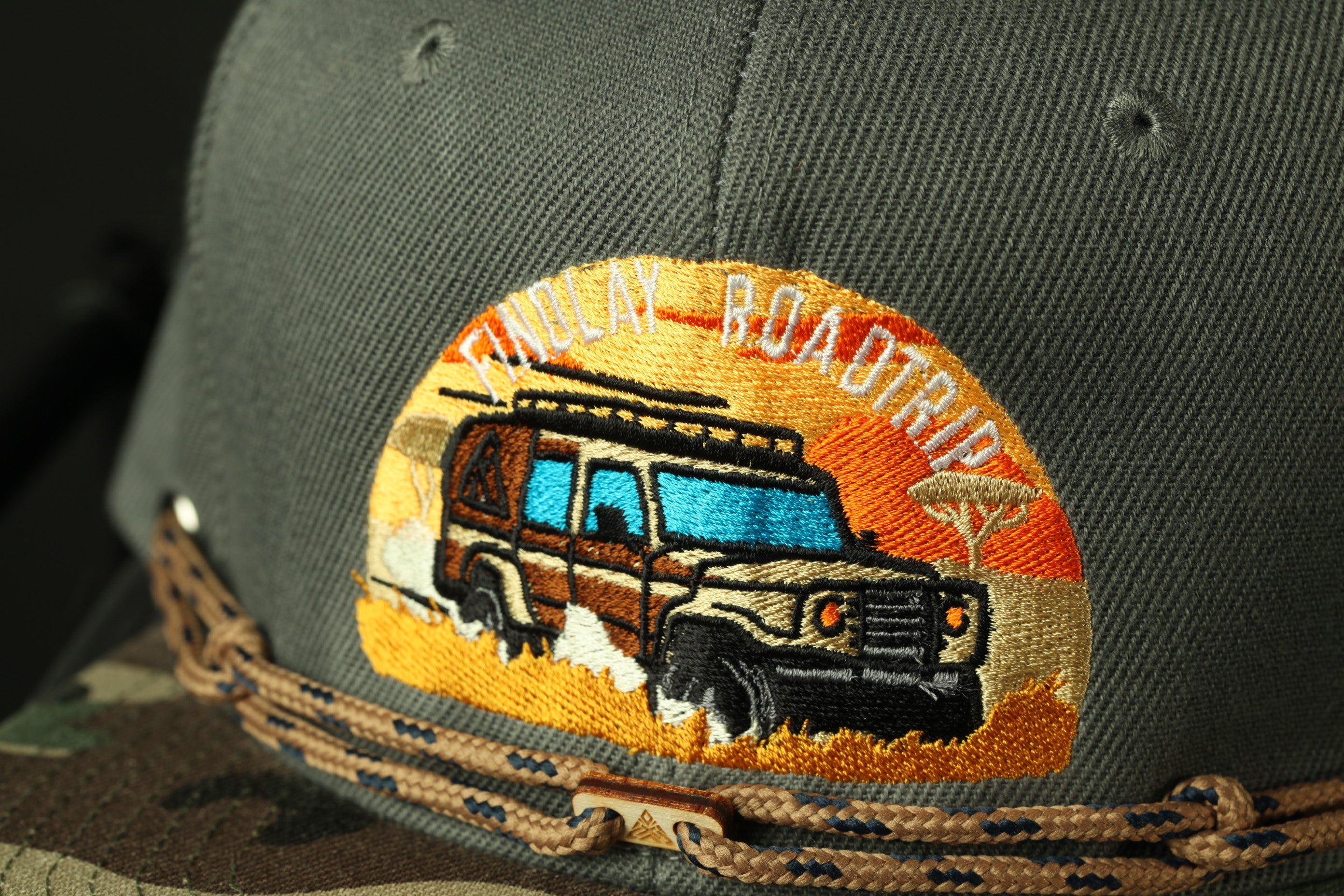 A close-up of a gray baseball cap with an embroidered design depicting a safari vehicle on a road.