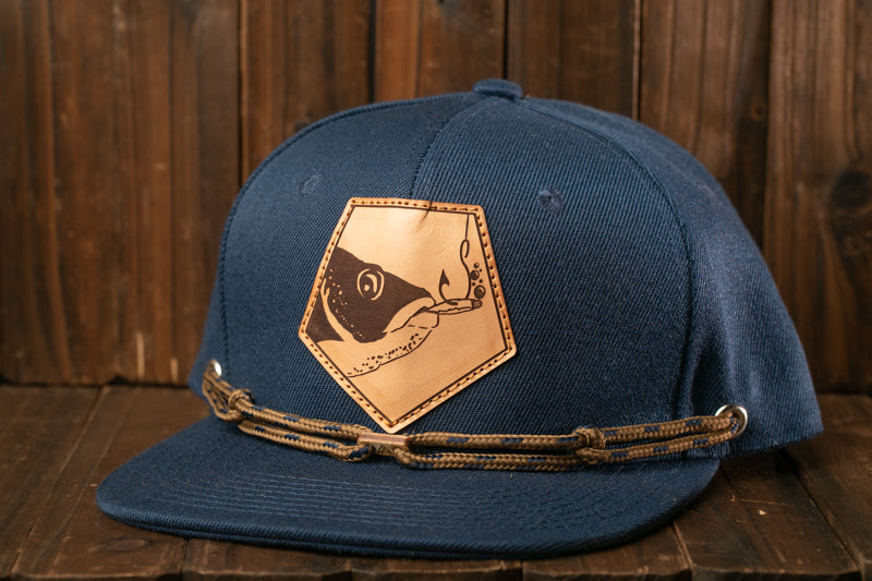 A navy blue baseball cap with a leather patch featuring a fish design, resting on a wooden surface.