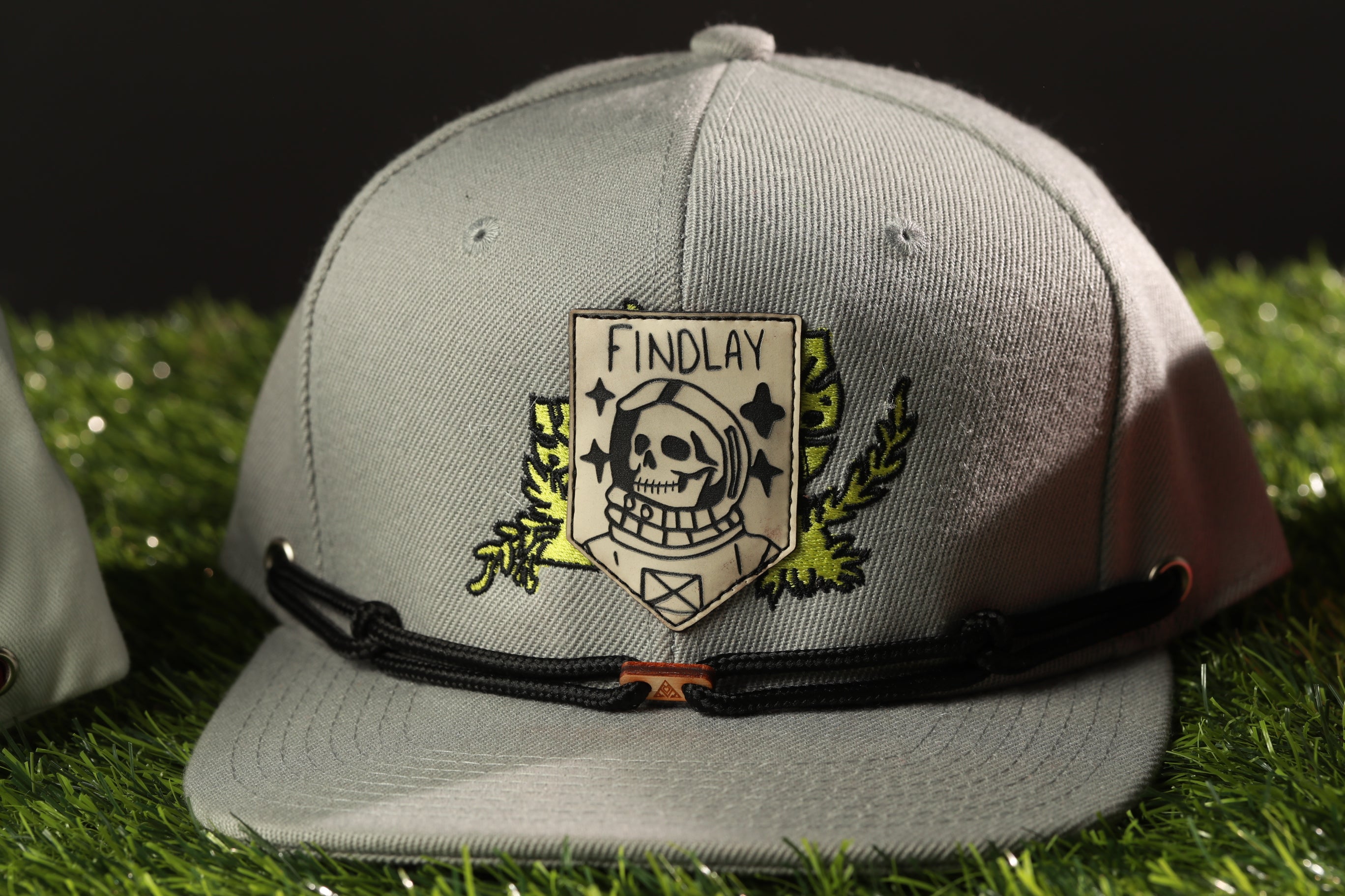 A gray baseball cap with a skull and crossbones design and the word "FINDA" printed on it, placed on a grassy surface.