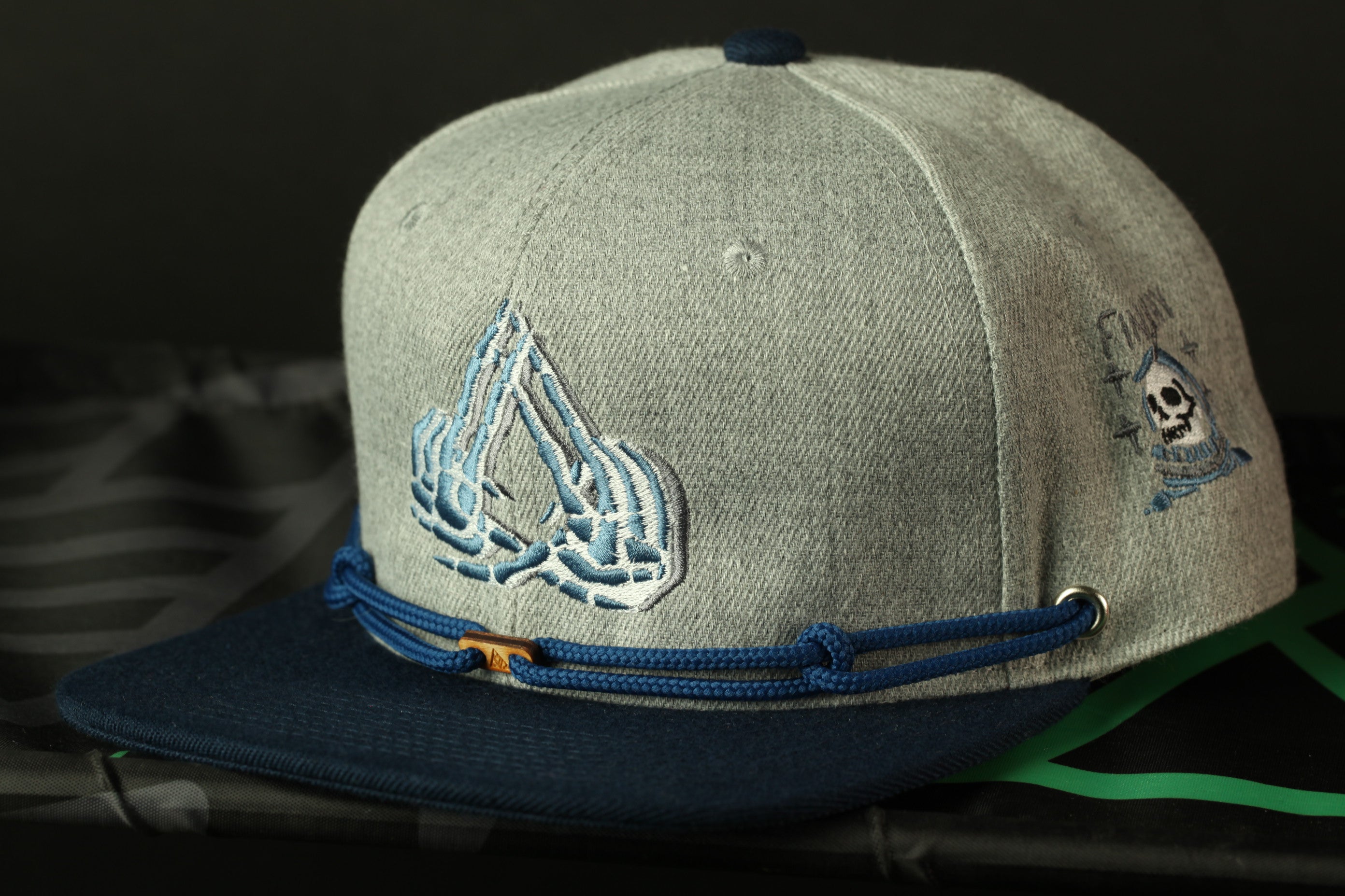 A gray baseball cap with a blue band and a skull design on the front, sitting on a black surface.