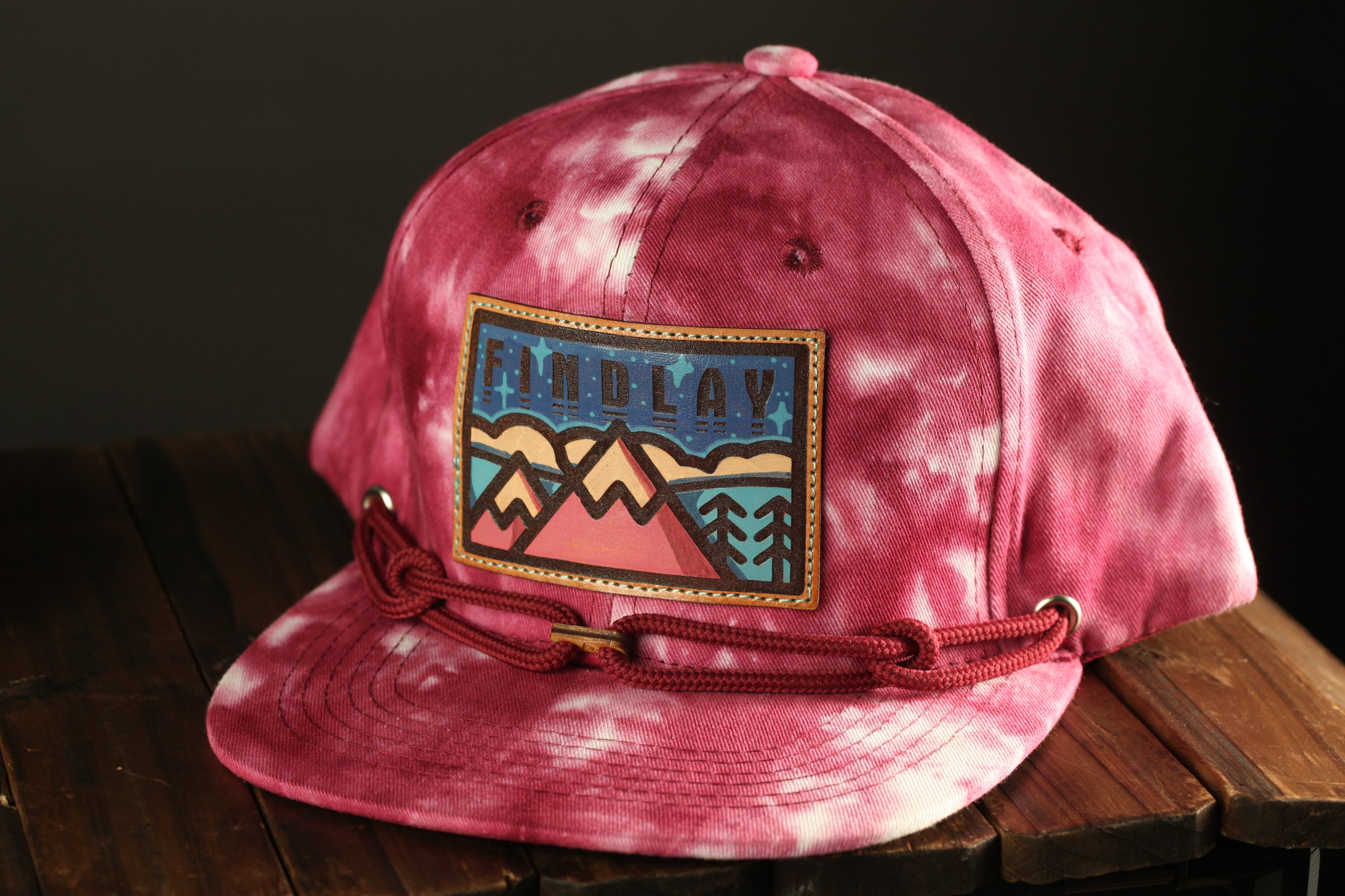 A red tie-dye baseball cap with a mountain landscape design on the front, sitting on a wooden surface.