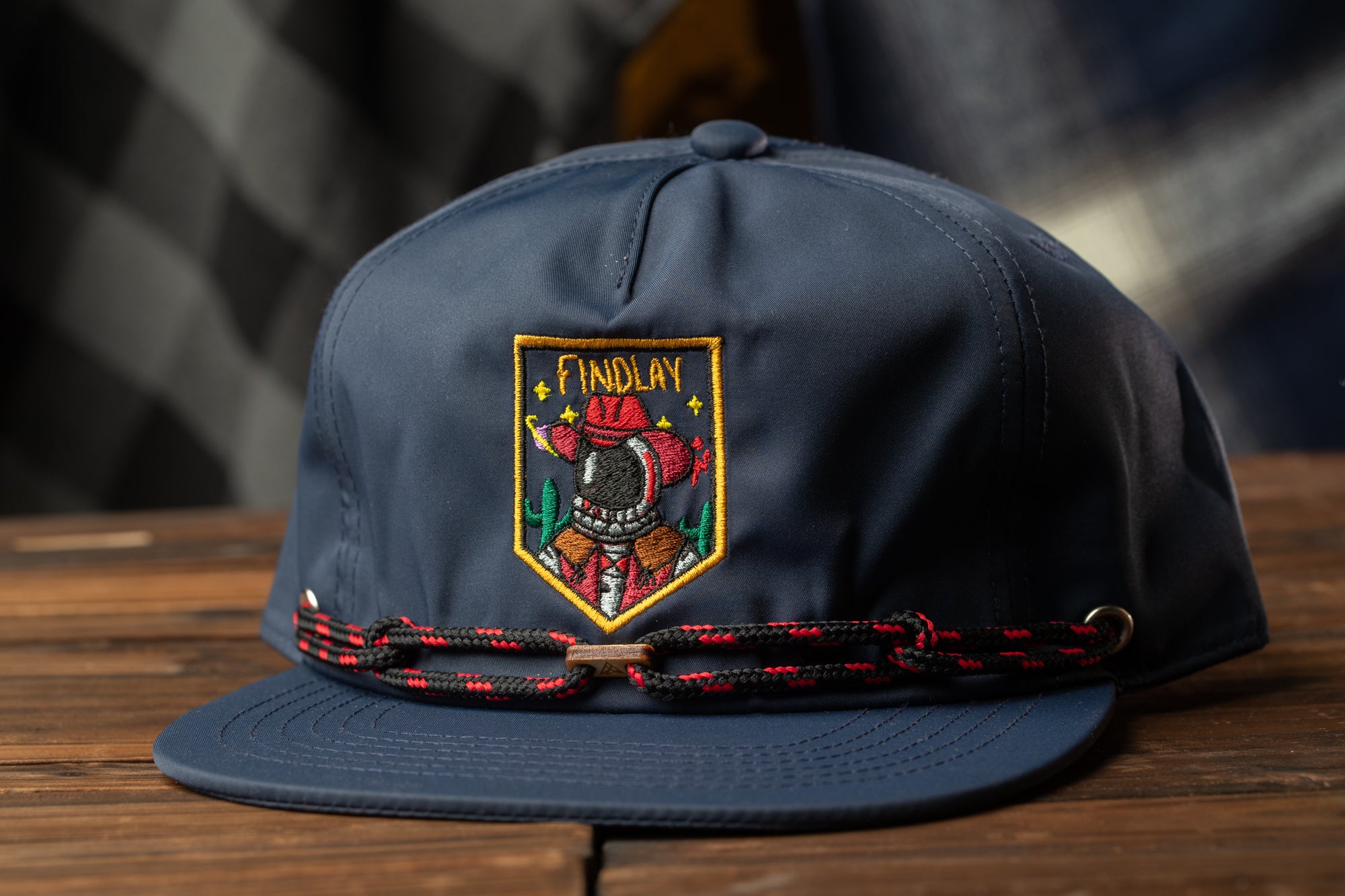 A navy blue baseball cap with a red and black braided band and an embroidered patch on the front, sitting on a wooden surface.