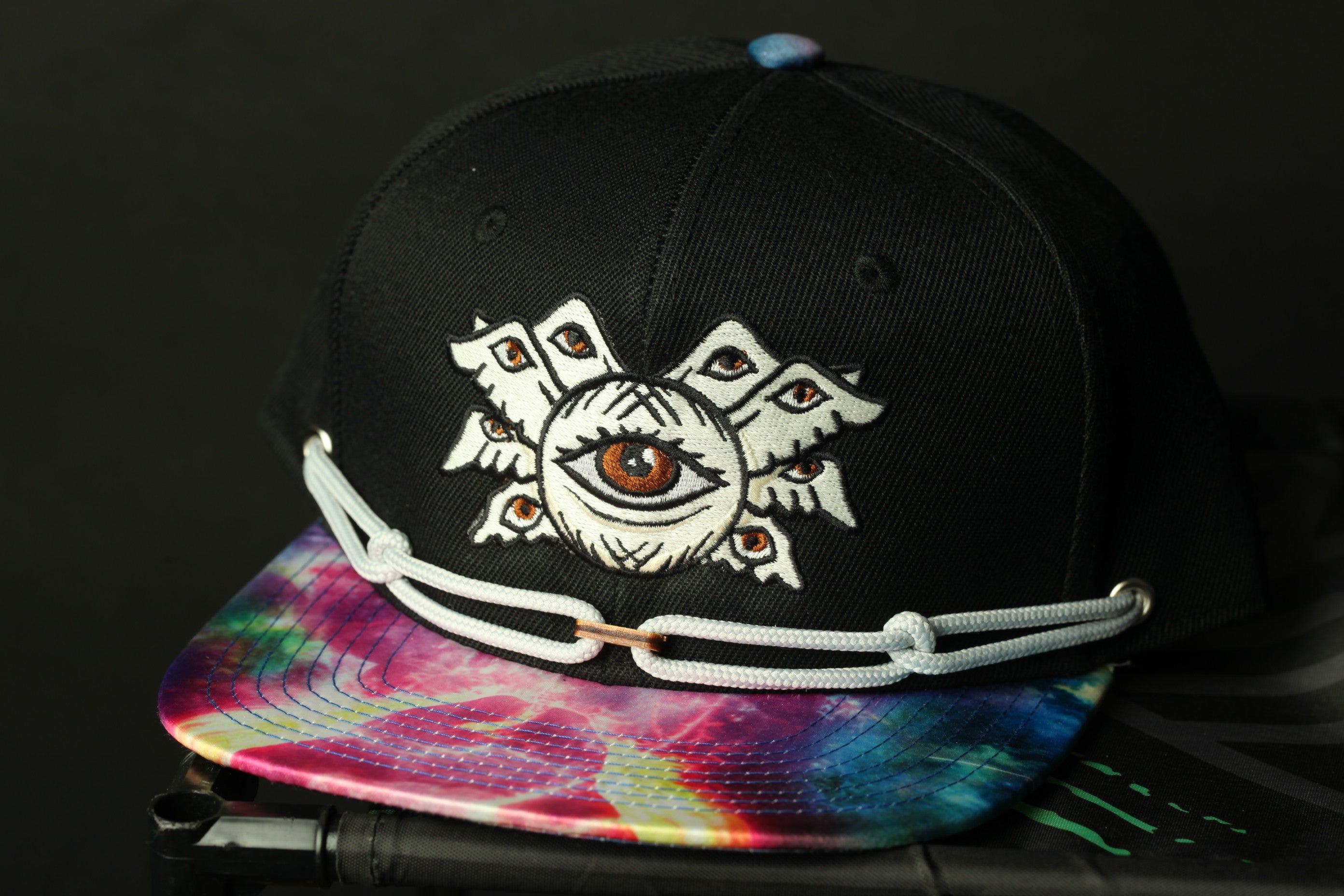 A black baseball cap with a colorful tie-dye pattern and an eye design on the front.
