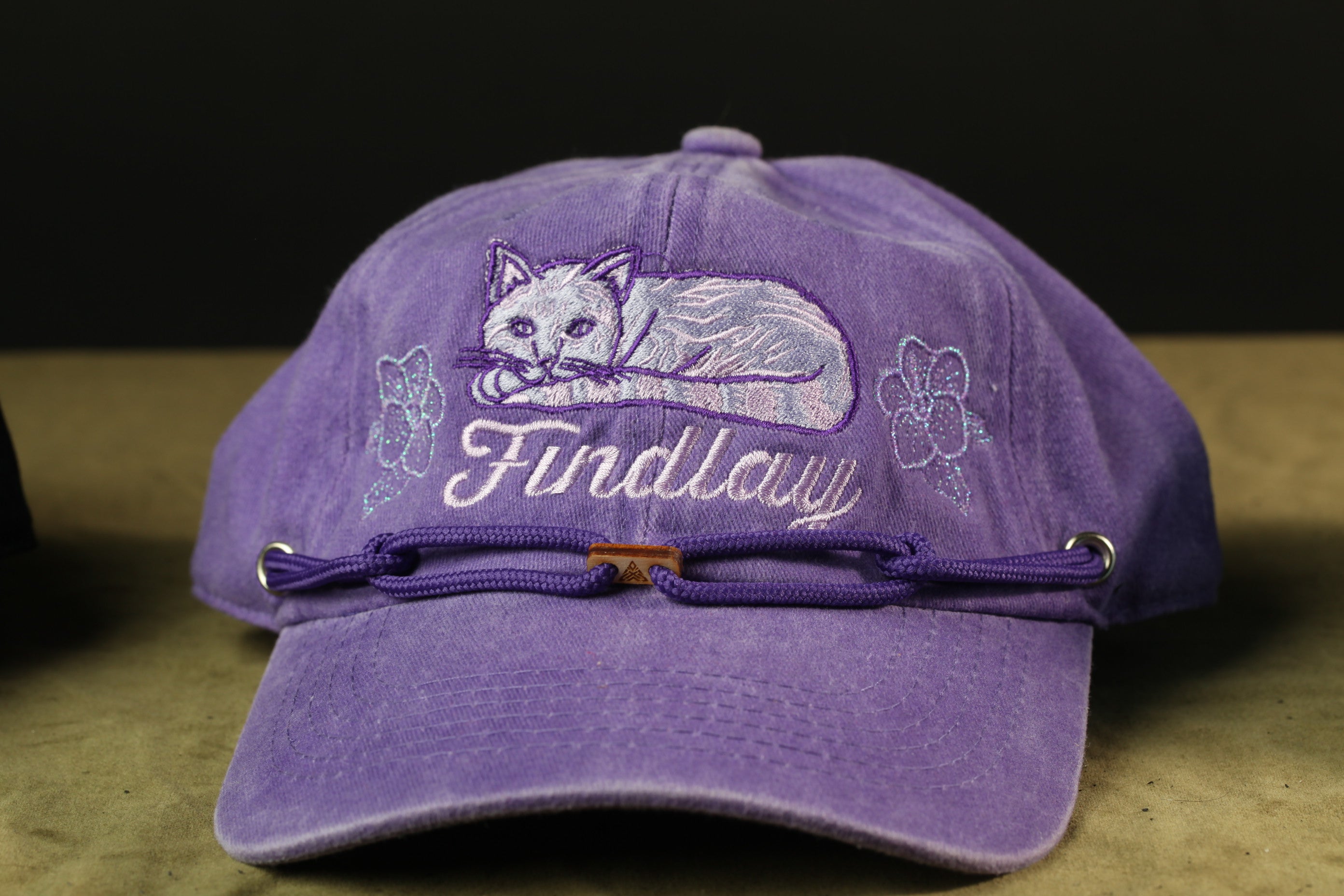 A purple baseball cap with a cat embroidered on it, and the word "Findlay" written on it.