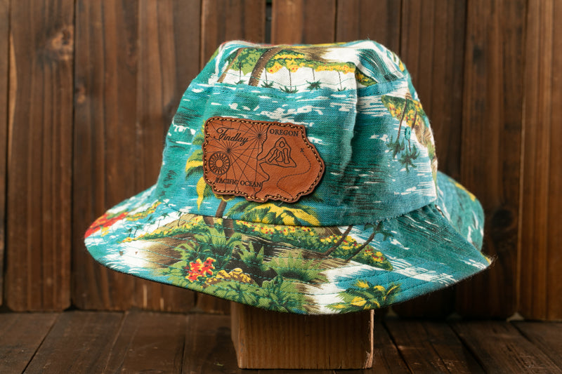A colorful Hawaiian-style bucket hat with a tropical print pattern and a leather patch on the front, displayed on a wooden surface.