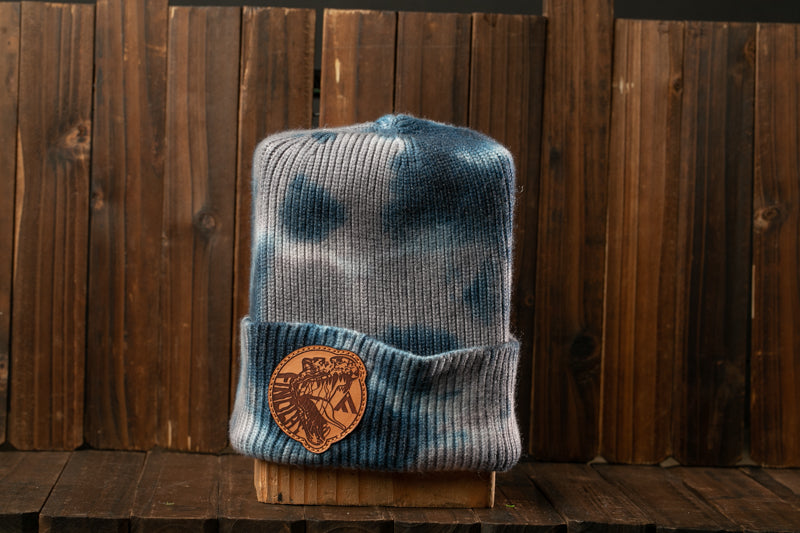 A gray and blue tie-dye beanie with a brown leather patch on the front, placed on a wooden surface against a wooden background.