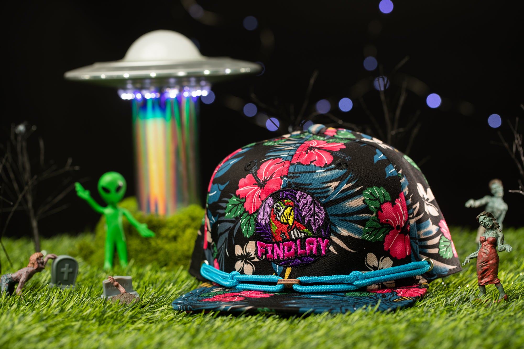 A colorful floral baseball cap with the word "FINDPLAY" on it, surrounded by various small toy figures and a large alien spacecraft in a grassy field.
