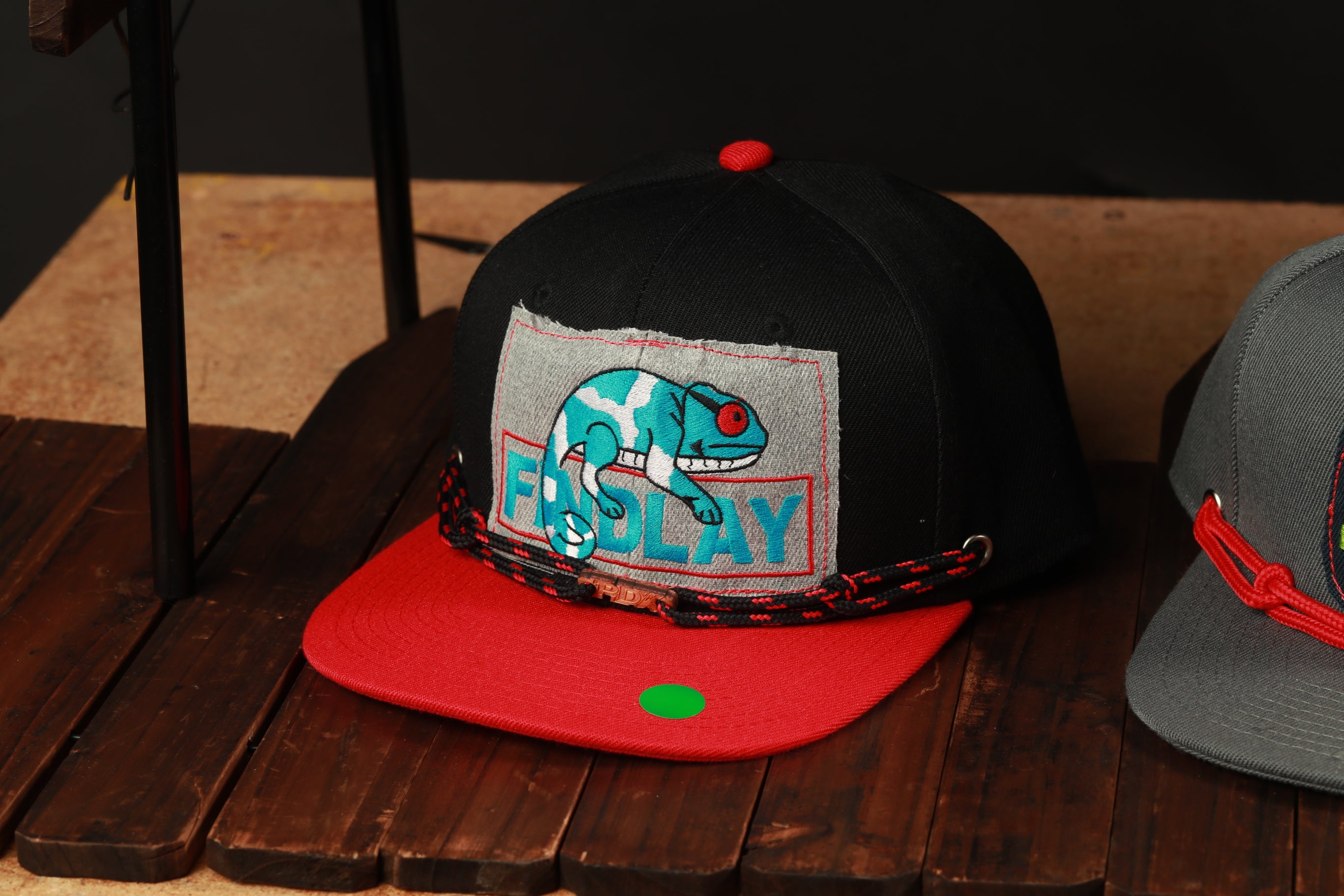 A black baseball cap with a red brim and a graphic of a chameleon is displayed on a wooden surface.