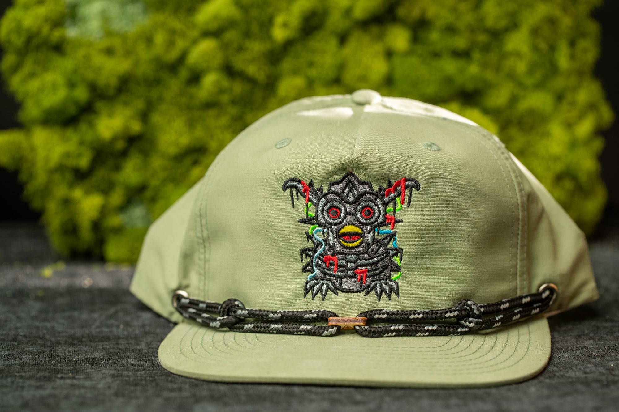 A beige baseball cap with a colorful, abstract embroidered design on the front, featuring a stylized, cartoonish face with red eyes and a red mouth.