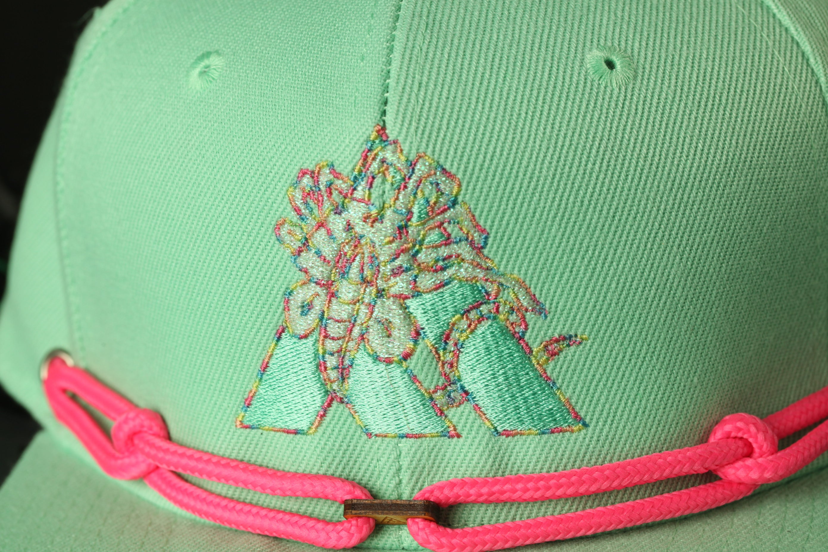 A close-up view of a green baseball cap with a pink braided strap and a colorful embroidered logo on the front.