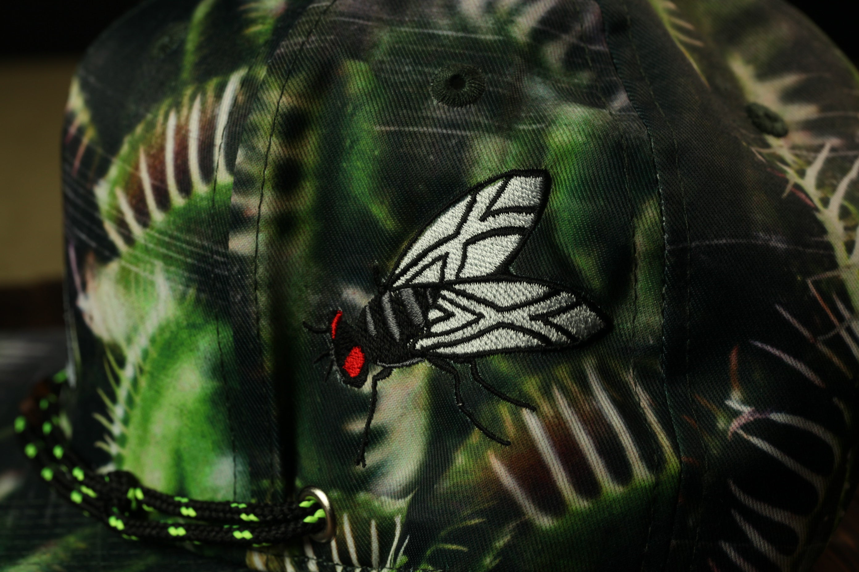 A close-up view of a camouflage-patterned baseball cap with a detailed embroidered design of a fly on the front.