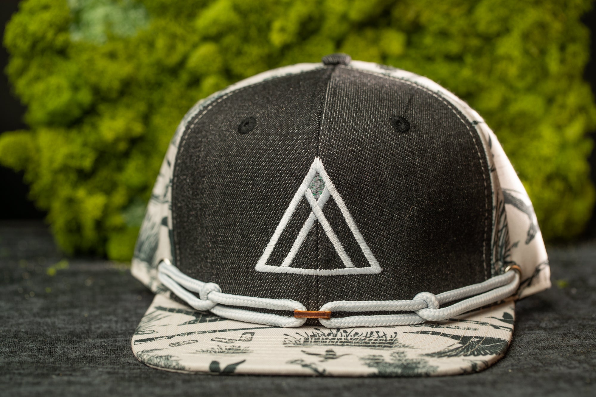 A black and white baseball cap with a white triangle logo on the front, placed on a gray surface against a background of green moss.