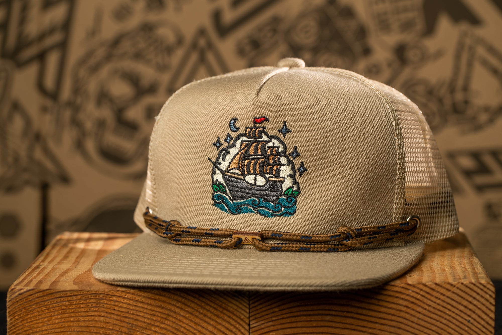A beige baseball cap with a detailed embroidered design of a ship on the front, sitting on a wooden surface against a blurred background.