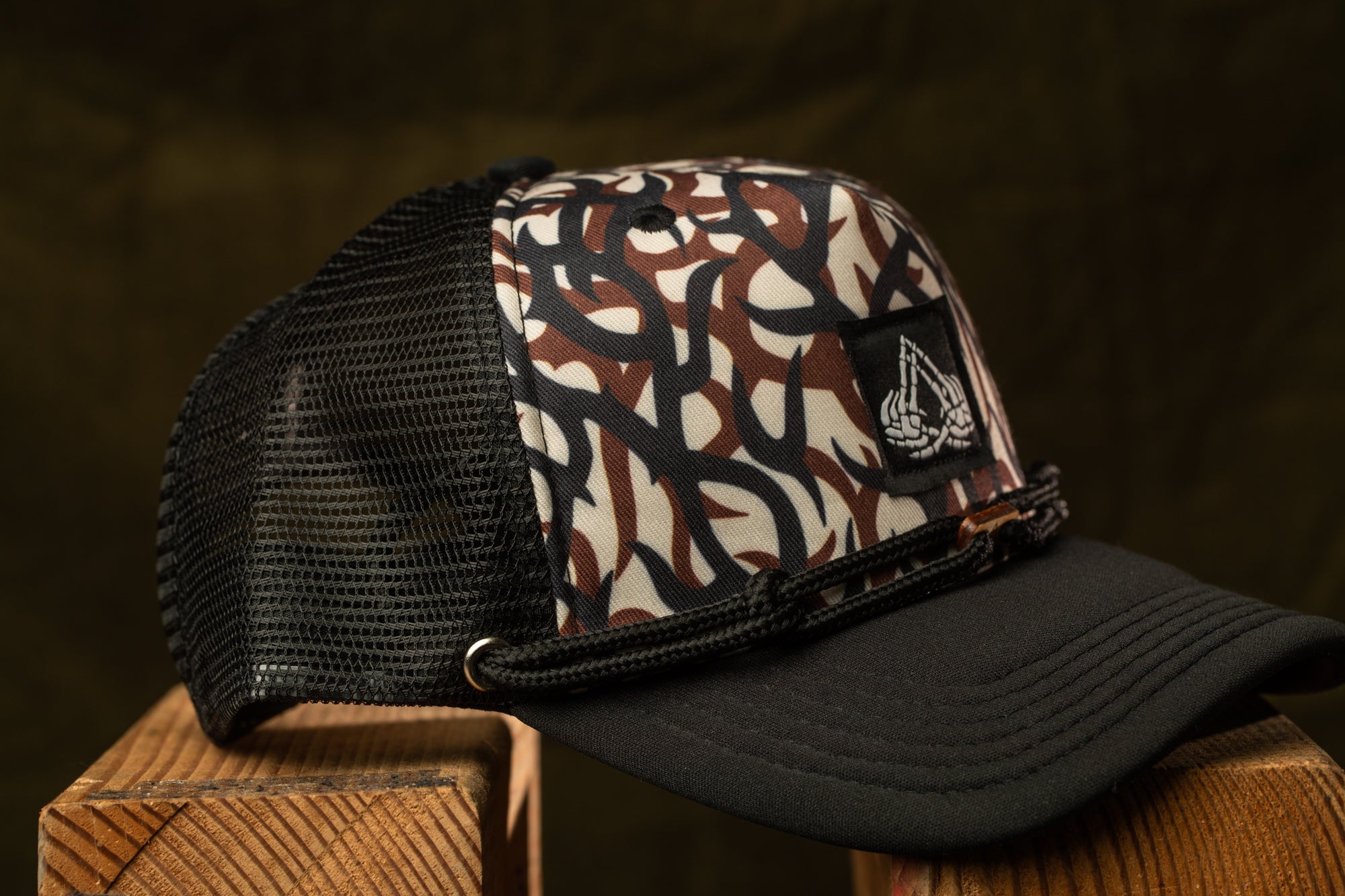 A black and brown camouflage baseball cap with a mesh panel and a logo on the front, resting on a wooden surface.