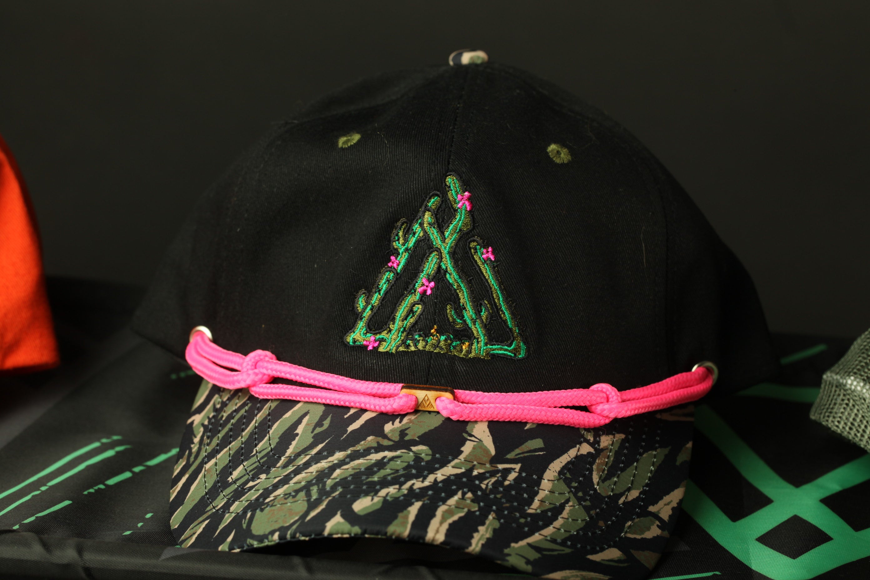 A black baseball cap with a pink braided strap and a green floral embroidered logo on the front.