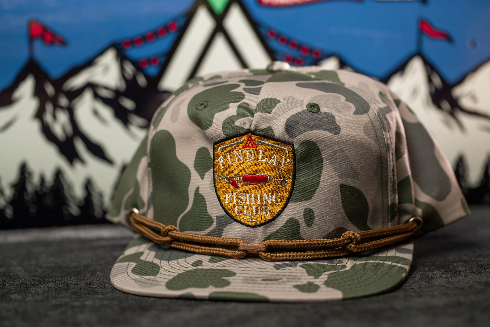 A camouflage baseball cap with a logo for the "Findlay Fishing Club" is displayed against a background of mountains and flags.