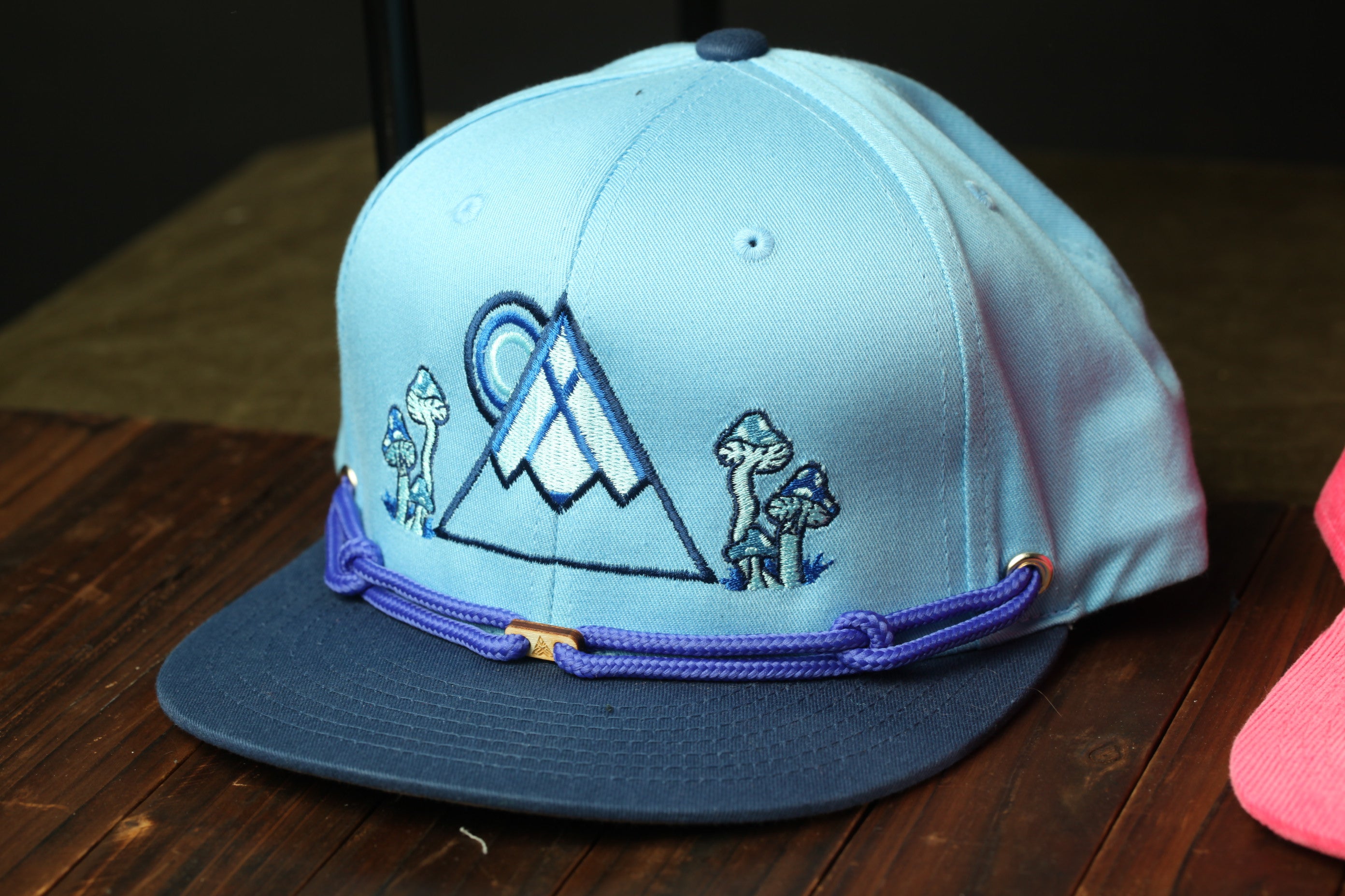 A blue baseball cap with a mountain and mushroom design, sitting on a wooden surface.