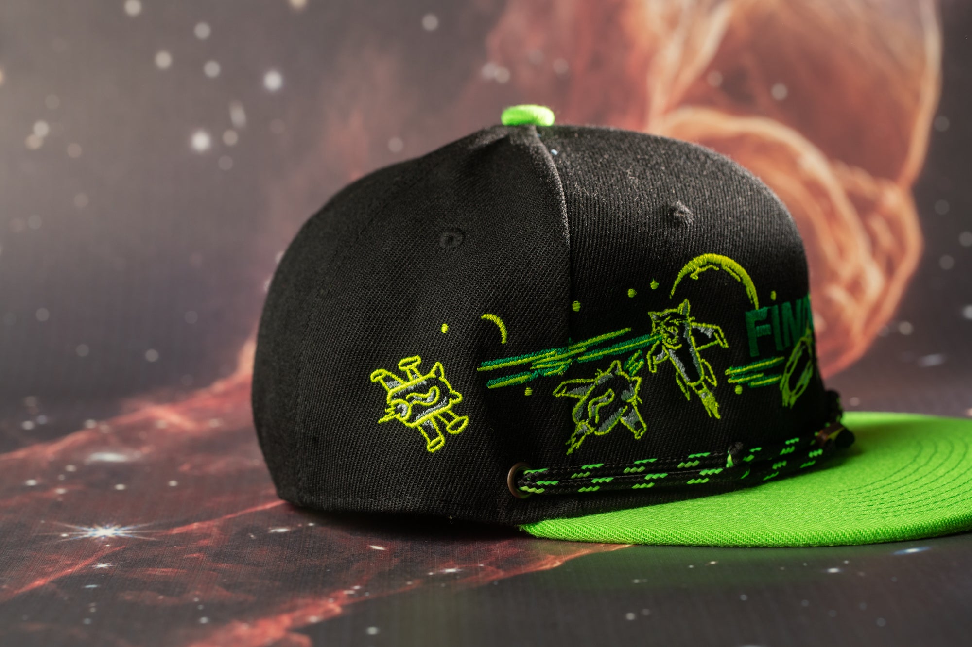 A black baseball cap with green and yellow designs, including a small alien-like creature, is shown against a dark background with a red and orange gradient.