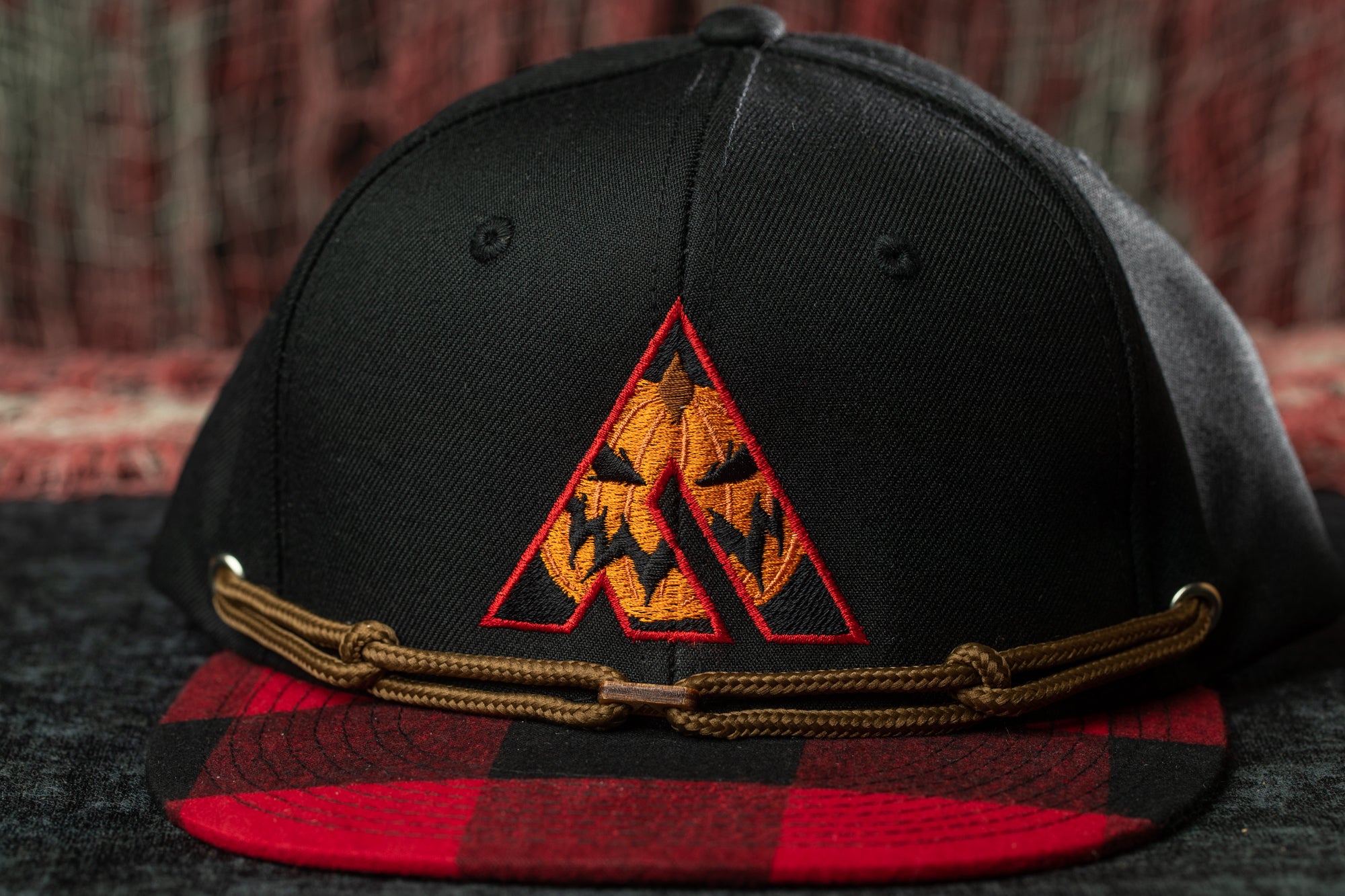 A black baseball cap with a red and black plaid pattern and a pumpkin-themed embroidered logo on the front.