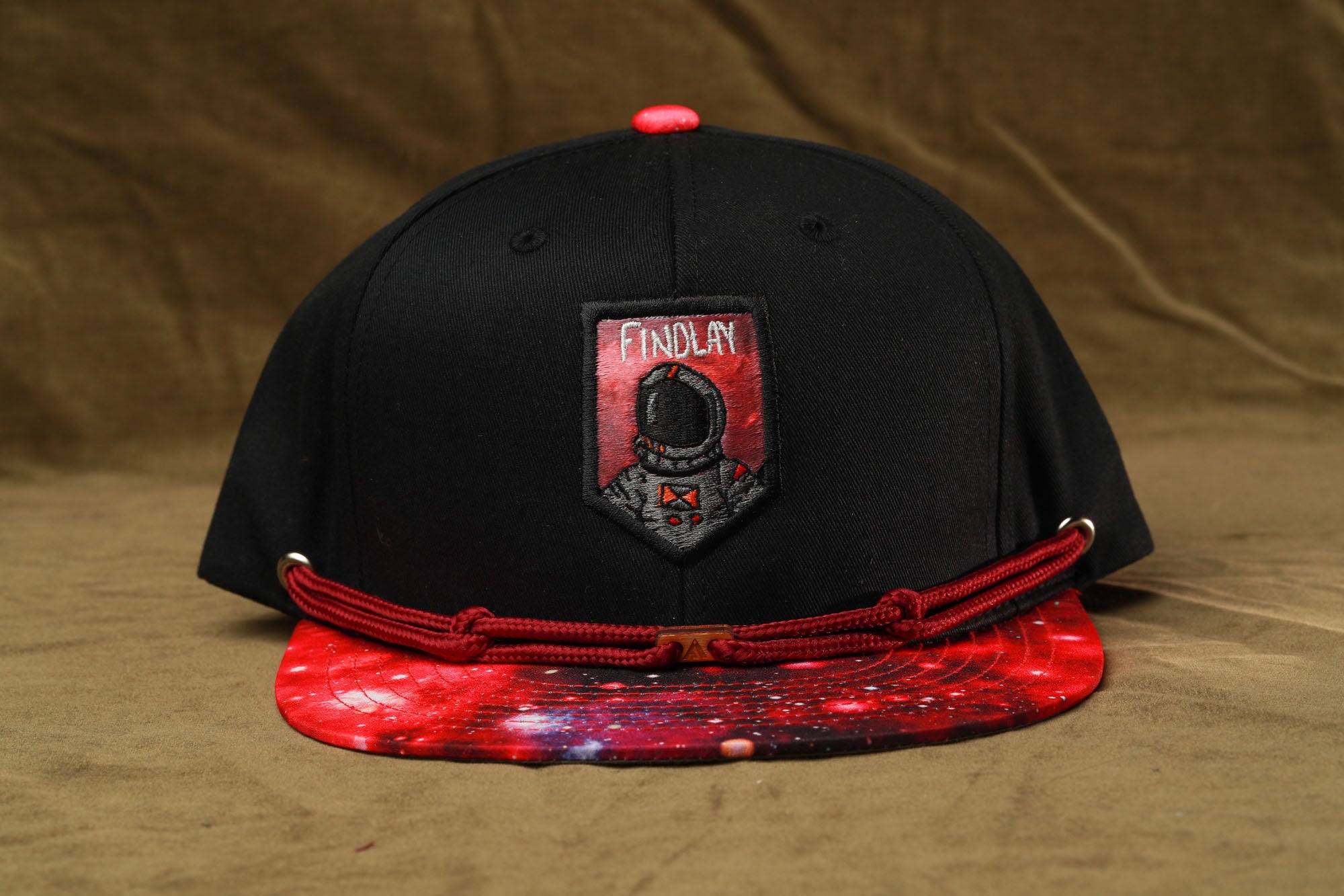 A black baseball cap with a red and white galaxy print on the front, featuring a patch with the text "FINDA" and an image of an astronaut.